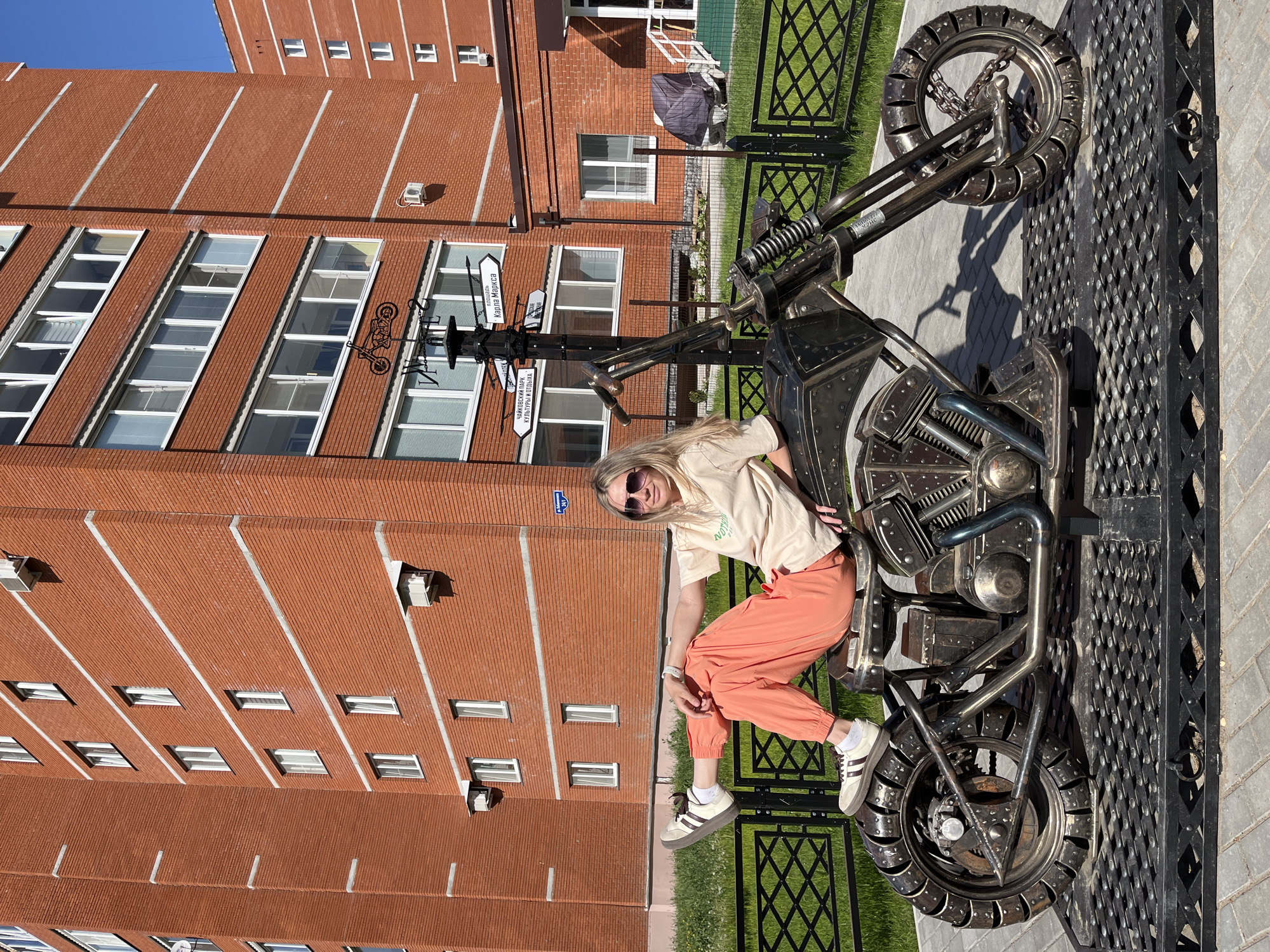 A fair-skinned female is sitting on a metal motorcycle sculpture in Chaikovsky, Russia. She has blonde hair and is wearing sunglasses, a light-colored t-shirt, and orange pants with white and black sneakers. She is positioned on the motorcycle as if she is riding it, with her legs slightly bent and her hands resting on the bike. The motorcycle is made entirely of metal parts and has a vintage or steampunk appearance. It is placed on a metal grate. Behind the motorcycle is a brick building with multiple windows and vertical white accents. A signpost with directional signs is visible near the motorcycle. A black metal fence is also in the background. The scene is well-lit and appears to be outdoors during the day.
