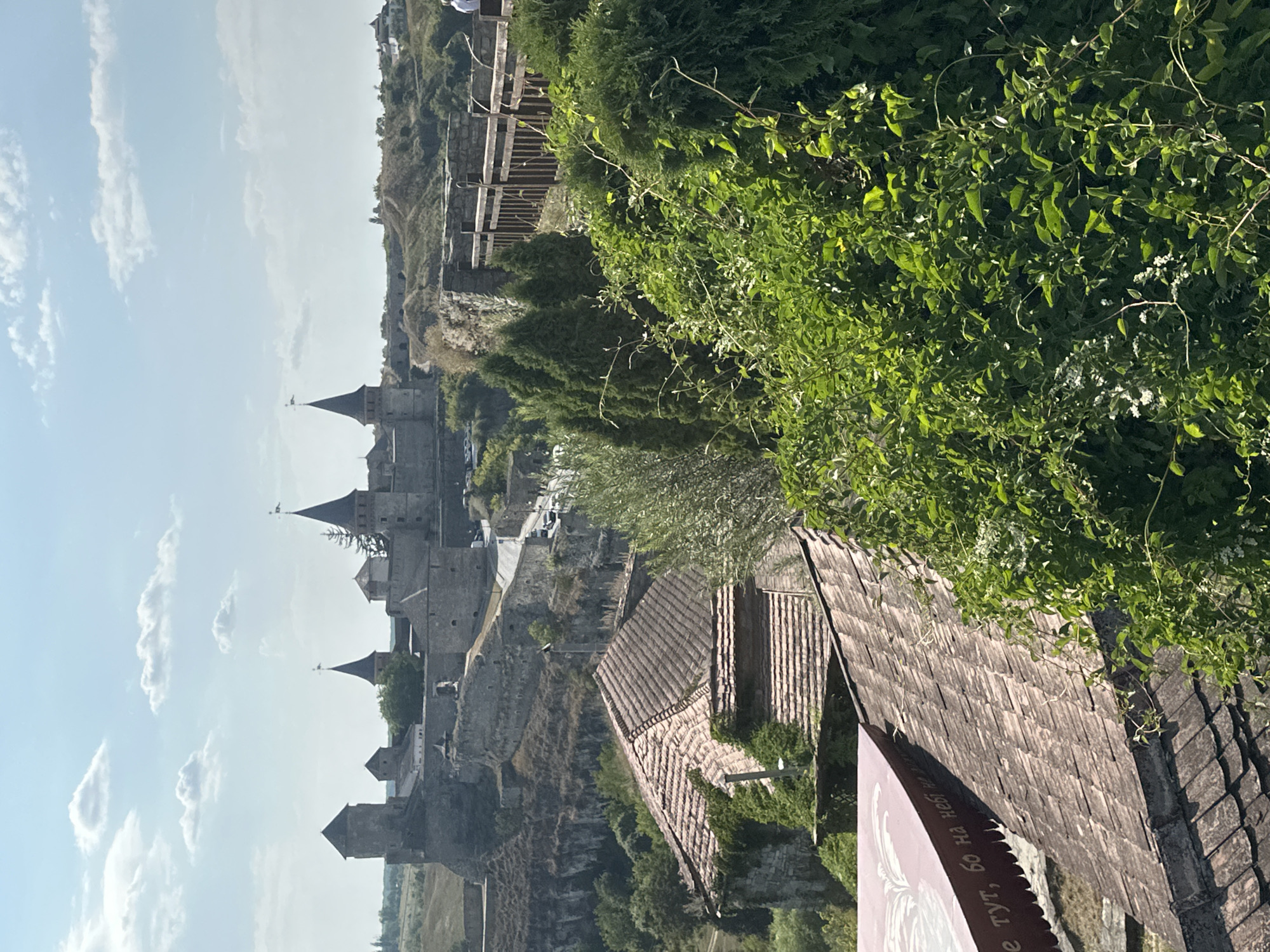 A vertical, slightly low-angle, full shot captures the Kamianets-Podilskyi Castle in Kamianets-Podilskyi, Ukraine, under a bright, partially cloudy sky. The castle, constructed of stone with conical towers and crenellations, sits atop a rocky hill. A road winds its way up to the castle, with cars visible in the distance. In the foreground, several older buildings with tiled roofs are partially overgrown with ivy and other greenery. A wooden fence runs along the top of a stone wall, overlooking the castle and the surrounding landscape. Lush green bushes and trees dominate the right side of the frame, partially obscuring the view of the castle. A small section of a red awning with Cyrillic text is visible in the lower left corner. The overall impression is one of a historic fortress situated amidst a verdant, hilly terrain.