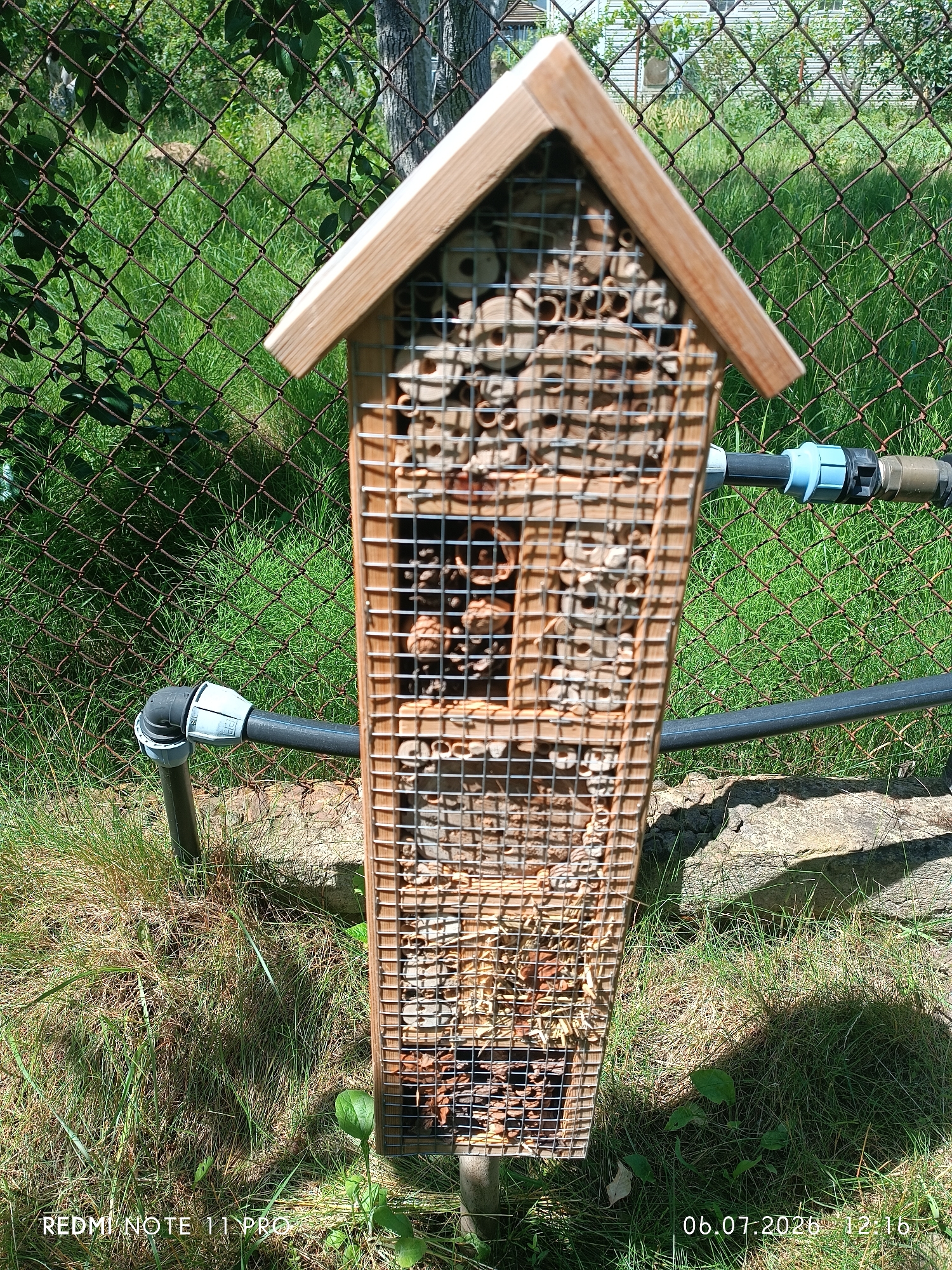 In a garden environment, a wooden insect hotel stands vertically. The hotel is constructed from natural materials such as hollow reeds, wood, and pine cones, and is covered by a wire mesh. A black hose with a blue connector runs horizontally behind the insect hotel. The ground is covered with grass, and in the background, a wire fence is visible. The image was captured on July 6, 2020, at 12:16 PM. The specific location is Vita-Poshtova, Ukraine.