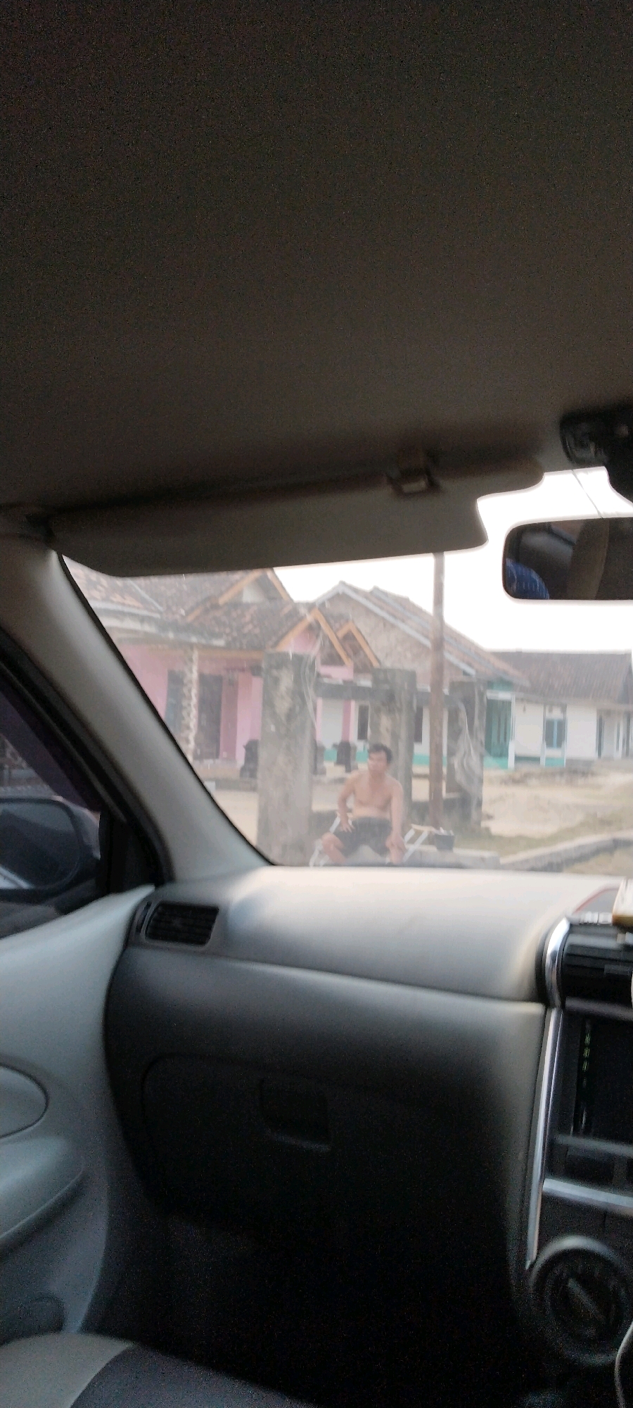 The image, taken from inside a vehicle, shows a man without a shirt sitting on the ground in front of some houses. The man is wearing dark shorts and appears to be looking forward. The houses in the background have pink and white facades with brown roofs. The scene appears to be a residential area in Tanjung Ratu Ilir, Indonesia, likely a rural or suburban setting given the modest houses and the presence of dirt or unpaved road. The lighting suggests it could be daytime, possibly late afternoon. The foreground is dominated by the interior of the car, including the dashboard, a side mirror reflecting the sky, and part of the car's roof lining. The overall image is slightly out of focus, particularly the background.