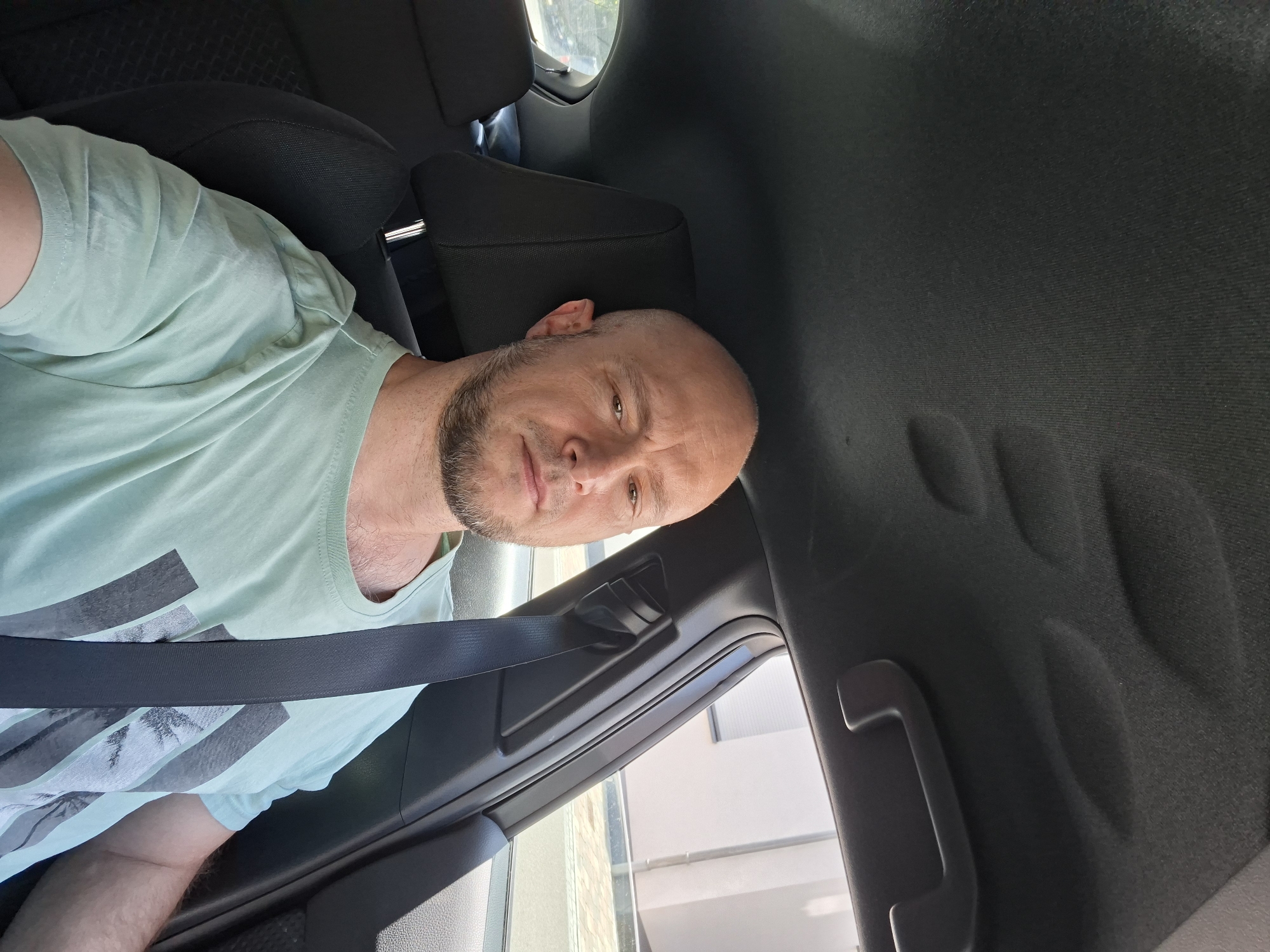 The image shows a man in the driver's seat of a car. He is bald with a beard and is wearing a light green t-shirt with a graphic print. The car's interior, including the headliner and seatbelt, is visible. The background outside the car shows a building and a street. The man is looking directly at the camera. The image appears to be a selfie taken inside the car. There is no indication of the location being Baden-Baden, Germany.