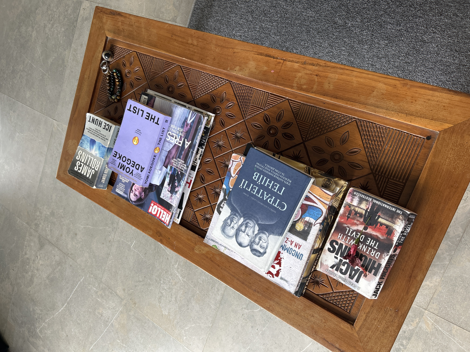 This is a high-angle, full shot of a wooden coffee table in Tembuku, Indonesia, with several books and jewelry displayed on it. The table has an intricately carved surface with floral and geometric patterns.
On the table, from top to bottom, are several books. The topmost books are two copies of "Ice Hunt" by James Rollins. Below these are two magazines and a book titled "The List" by Yomi Adegboke. Further down, a book titled "Стратегії геніїв" (Strategies of Geniuses) in Ukrainian is visible, with a black and white image of three men on its cover. Beneath that, a book with the title "UNCOMMON AN A-Z" is partially visible. Finally, the bottommost book is "Drink with the Devil" by Jack Higgins, featuring an image of London's skyline and a red flower placed on top. A bracelet made of green and brown beads and two rings are placed near the books. The table is situated on a textured grey tiled floor, with a dark grey rug partially visible on the right side. The lighting suggests it is daytime, with natural light illuminating the scene.