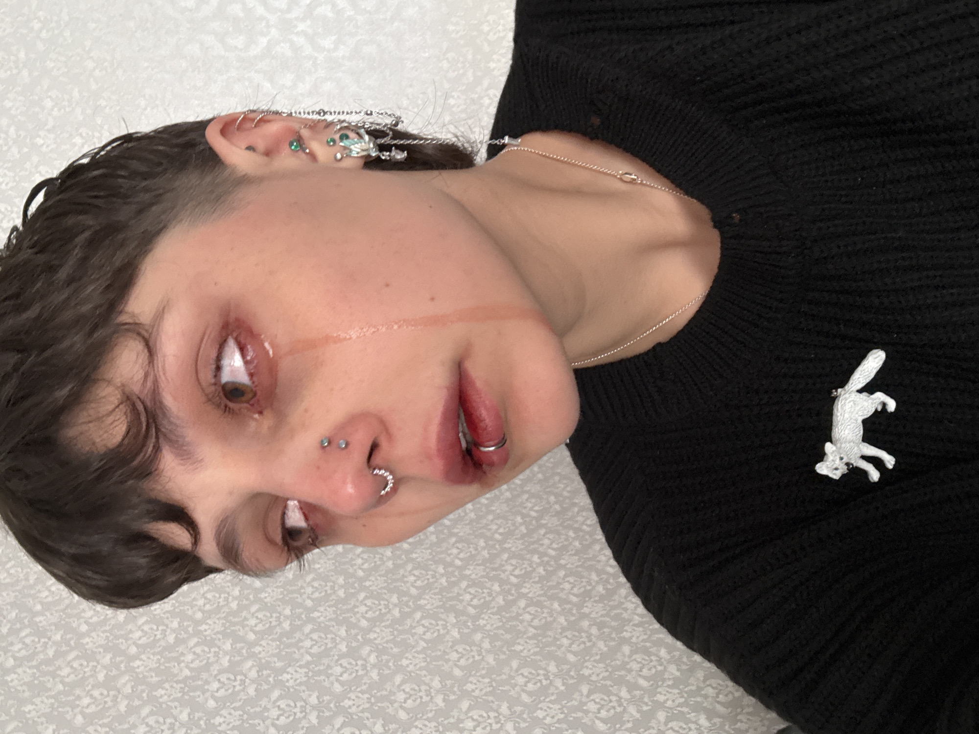 This is a close-up, rotated portrait of a person, positioned with their head on the left and body extending to the right. The individual has short, dark, curly hair and striking brown eyes. They wear a black knitted sweater, adorned with a small, white fox-shaped brooch on the right side of their chest.
The person exhibits a significant number of silver-toned facial and ear piercings, including multiple studs, hoops, and chains on their ears, some embellished with small green gemstones. A double nostril piercing, a horseshoe-shaped septum piercing, and a silver hoop on their lower lip are also prominent. A thin silver chain necklace is visible around their neck.
A notable detail is the presence of liquid streaks, appearing as tears or a stylized artistic effect, running from the inner corner of their visible eye down across their cheek. The person's eyes are wide open, and their mouth is slightly ajar. The background is a light-colored surface with a subtle, repeating pattern, indicating an indoor setting. The overall aesthetic is expressive and leans towards an alternative style, possibly conveying a sense of vulnerability or artistic drama. No text is visible in the image. The context for this media file is Saint Petersburg, Russia.