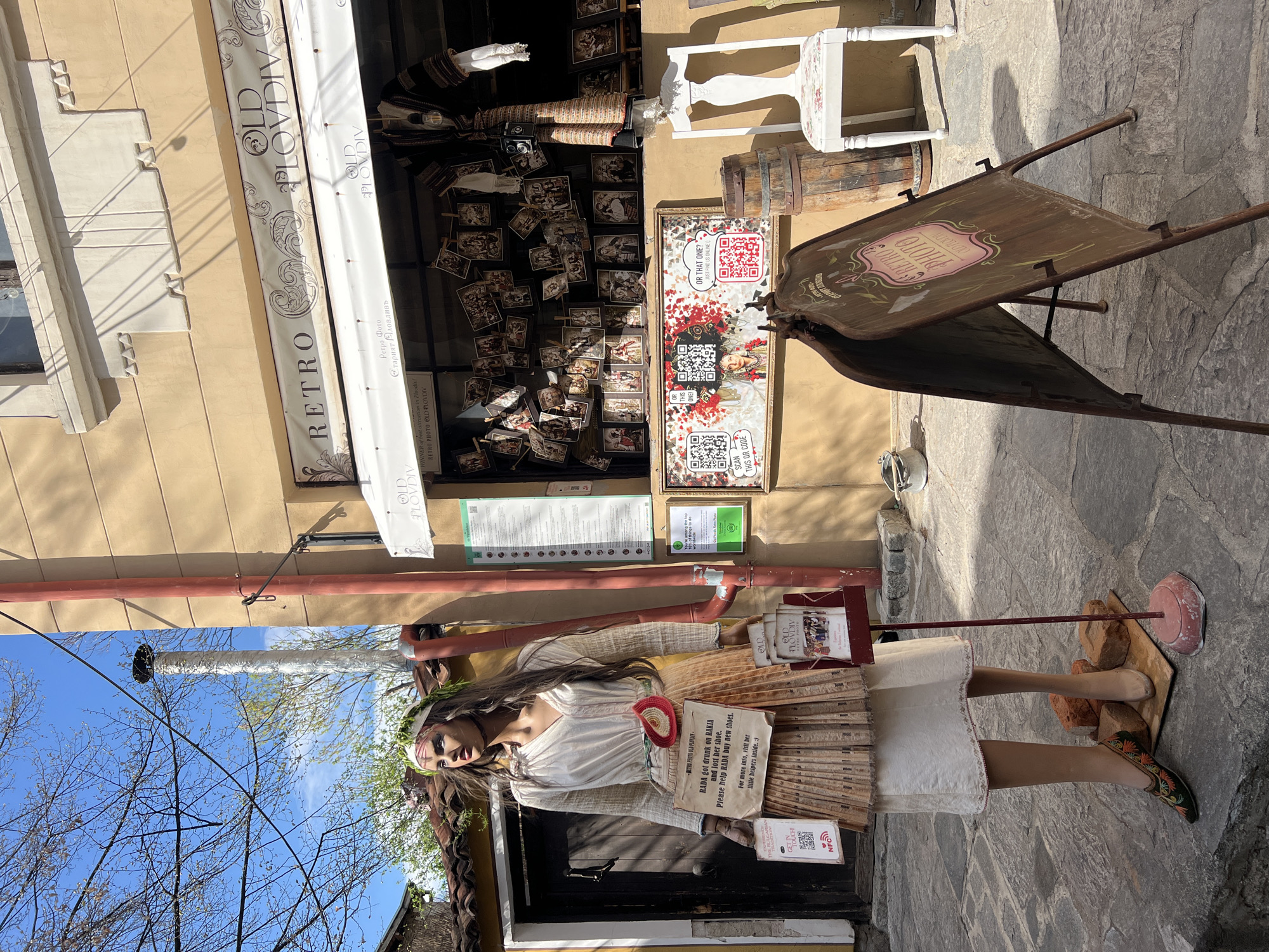 The image captures a street scene in Plovdiv, Bulgaria, with the "RETRO PHOTO OLD PLOVDIV" store prominently displayed. The storefront has a white awning with the shop's name in brown lettering. A mannequin dressed in traditional attire stands in front of the shop, holding signs and brochures, with one sign stating that "RADA got drunk on RAKIA and lost her shoe" and asking for help to buy new shoes. The shop window is filled with old photographs clipped onto strings, and a wooden barrel and chair are visible. A sandwich board-style sign advertising "RETRO PHOTO" is placed to the side. The ground is paved with stone, and a glimpse of tree branches and the sky is visible in the upper left corner. The image is rotated sideways.