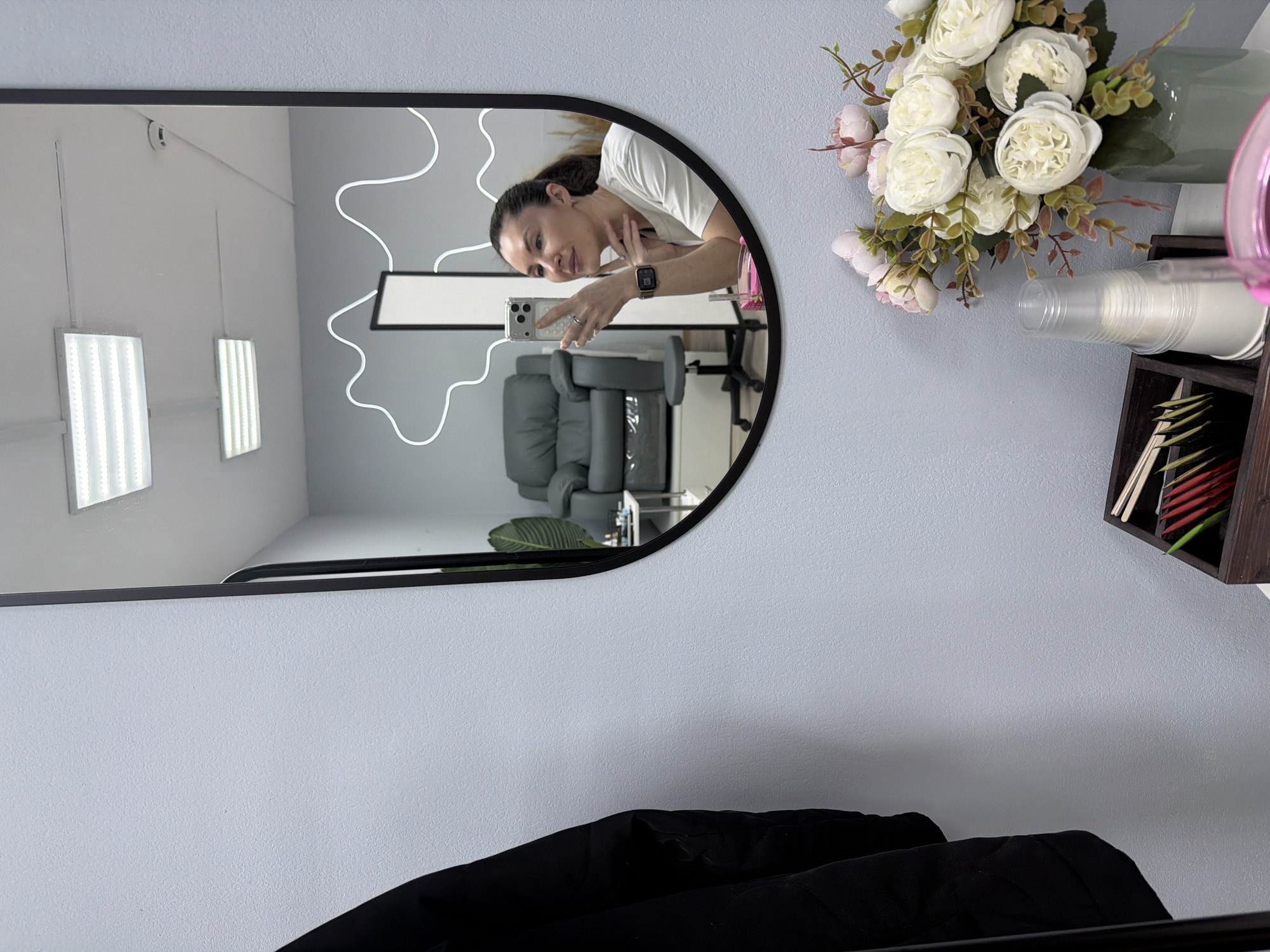 The image shows a mirror reflecting the interior of a business, potentially a beauty salon or spa, in what appears to be Perm, Russia. In the reflection, a woman is taking a selfie with her smartphone. She is wearing a white shirt and a smartwatch on her left wrist. The interior includes treatment chairs and a decorative light fixture on the wall. To the right of the mirror are flowers, disposable cups, and a small organizer with tea bags or similar items. At the bottom of the image, a black jacket or coat is visible, partially obscuring the wall below the mirror.