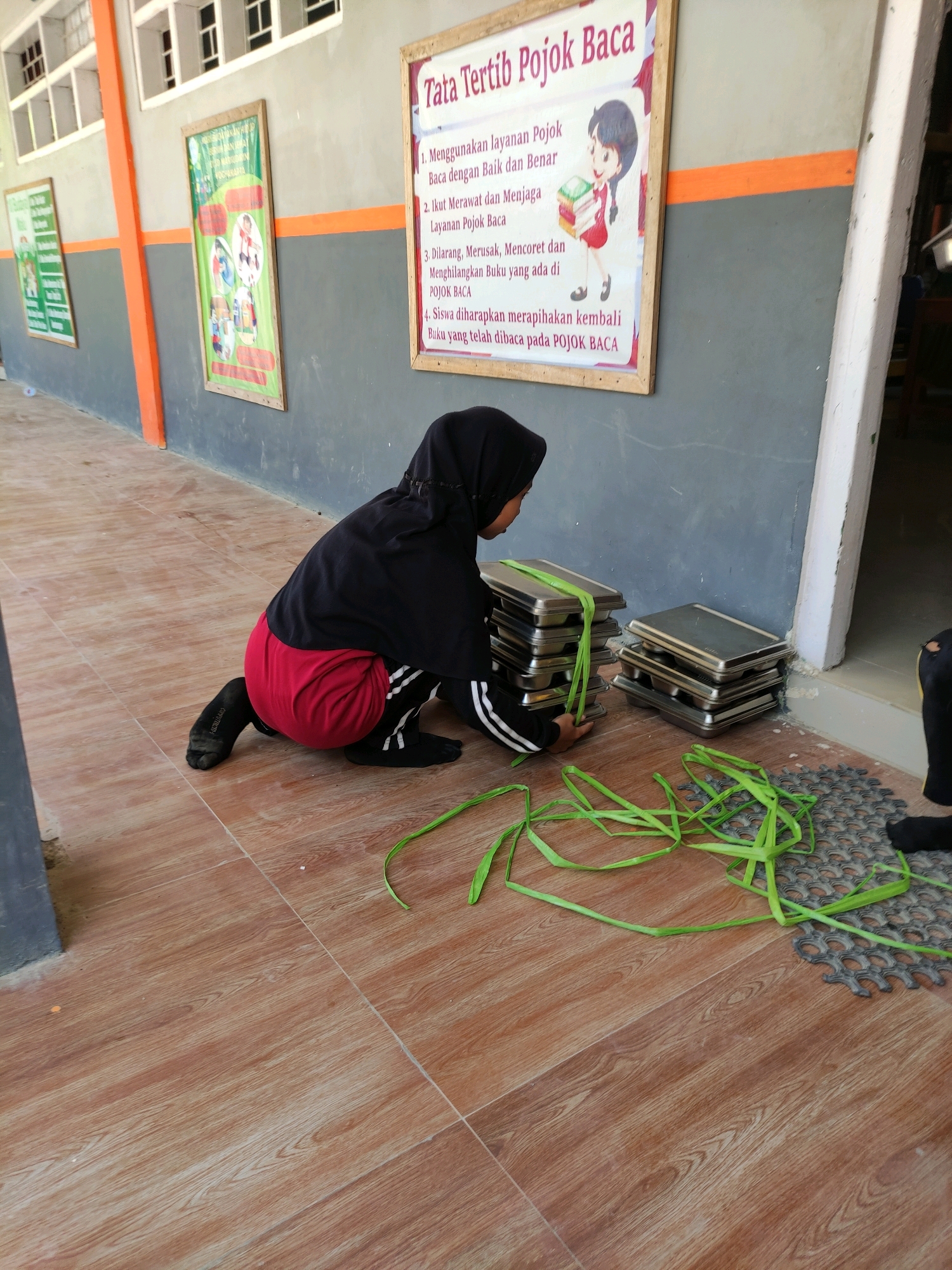 A young person wearing a black hijab, black long-sleeved shirt, red shorts, and black socks is kneeling on a tiled floor. The person is interacting with stacks of metal trays that are tied together with green plastic strips. The trays are arranged in two separate stacks. One stack is larger and to the left, while a smaller stack is to the right. Green plastic strips are scattered on the floor around the trays. In the background, there is a grey wall with an orange stripe. A framed poster with Indonesian text and an illustration of a cartoon character holding books is hanging on the wall. Another poster with text is visible further to the left. The flooring is composed of brown tiles with a wood grain pattern. The scene appears to be outdoors or in an open, covered area, possibly a school hallway or a communal space in Bulukumba, Indonesia, given the posters.