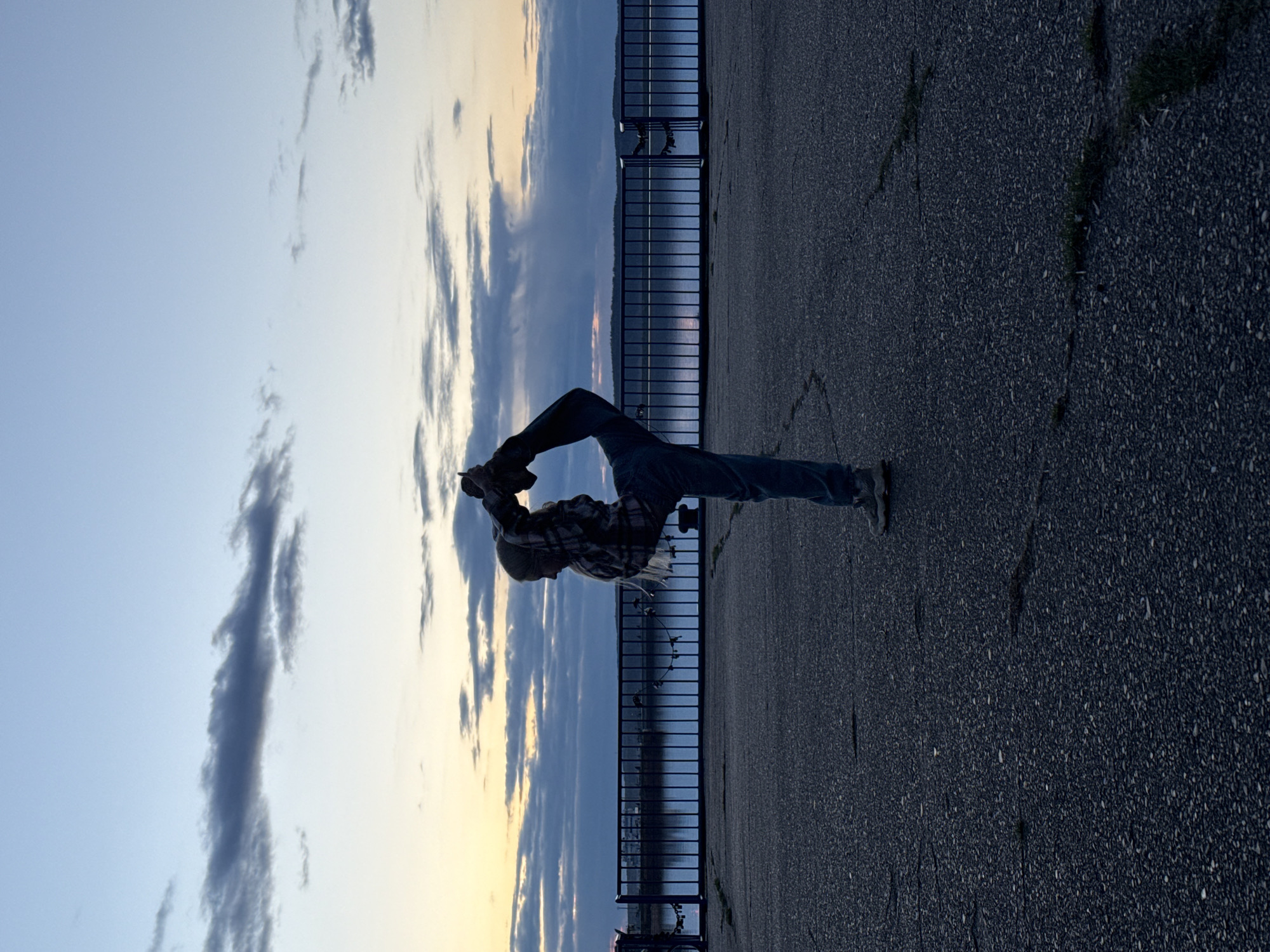 The image, when viewed in its correct upright orientation, portrays a solitary person standing on a paved embankment or promenade, gazing towards a vast body of water under a late afternoon or early evening sky. The sky is adorned with scattered clouds, displaying a gentle gradient from pale blue to deeper blues and hints of warm yellow and orange near the horizon, indicative of sunset or twilight.
In the center, a person is seen from a rear-three-quarter angle, dressed in a plaid shirt and blue jeans. They are actively holding up a camera or smartphone, presumably to photograph the expansive water and sky before them.
A dark, vertical-bar metal railing runs along the edge of the paved area, separating it from the tranquil water beyond. The water's surface subtly reflects the sky's soft colors. The ground in the foreground and beneath the person's feet is a dark, coarse-grained pavement, likely asphalt or concrete, showing some cracks and sparse patches of grass.
The scene conveys a sense of calm and appreciation for nature, capturing a moment of observation or documentation. This peaceful outdoor setting, with its specific light quality, points to a location such as a waterfront in Chaikovsky, Russia. No visible text is present in the image.