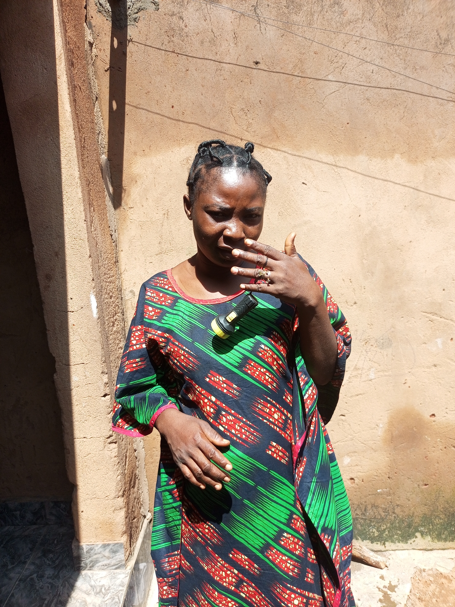 A medium shot captures a Black woman standing outdoors in front of a textured, tan-colored wall. The woman, facing slightly away from the camera and to the right, has her hair styled in multiple braids. Her right hand is raised to her face, with her fingers touching her mouth and nose, and she is wearing several rings on her fingers. Her left hand rests on her abdomen. She is wearing a colorful, flowing dress with a pattern of black, green, and red geometric shapes. A yellow and black flashlight is clipped to the neckline of her dress. The wall behind her has some visible cracking and texture. To the left of the woman, a dark, recessed doorway is visible, with tiled flooring in the foreground. Sunlight casts a shadow on the wall to the left of the woman.
