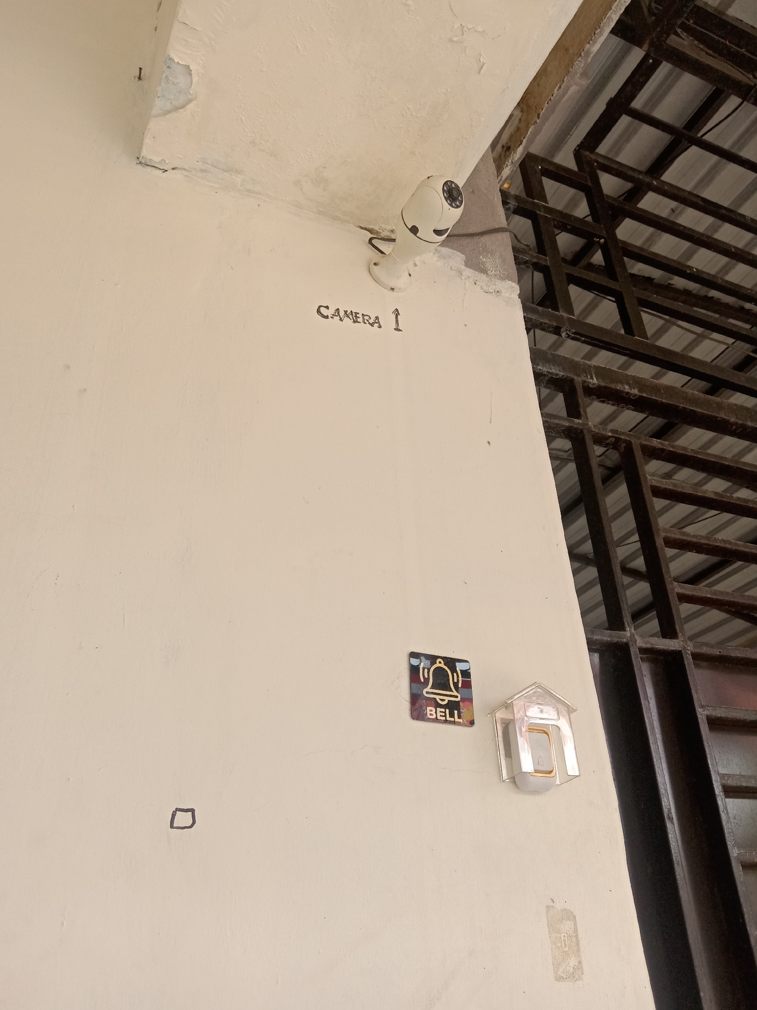 In Stiaji, Indonesia, a white wall displays a security camera labeled "CAMERA 1" near the ceiling. Below it, a doorbell with a small bell icon is affixed to the wall, accompanied by a separate sticker that also reads "BELL." A small, drawn square is visible on the lower left portion of the wall. The scene is partially obscured by a metal grate structure above and to the right of the wall.