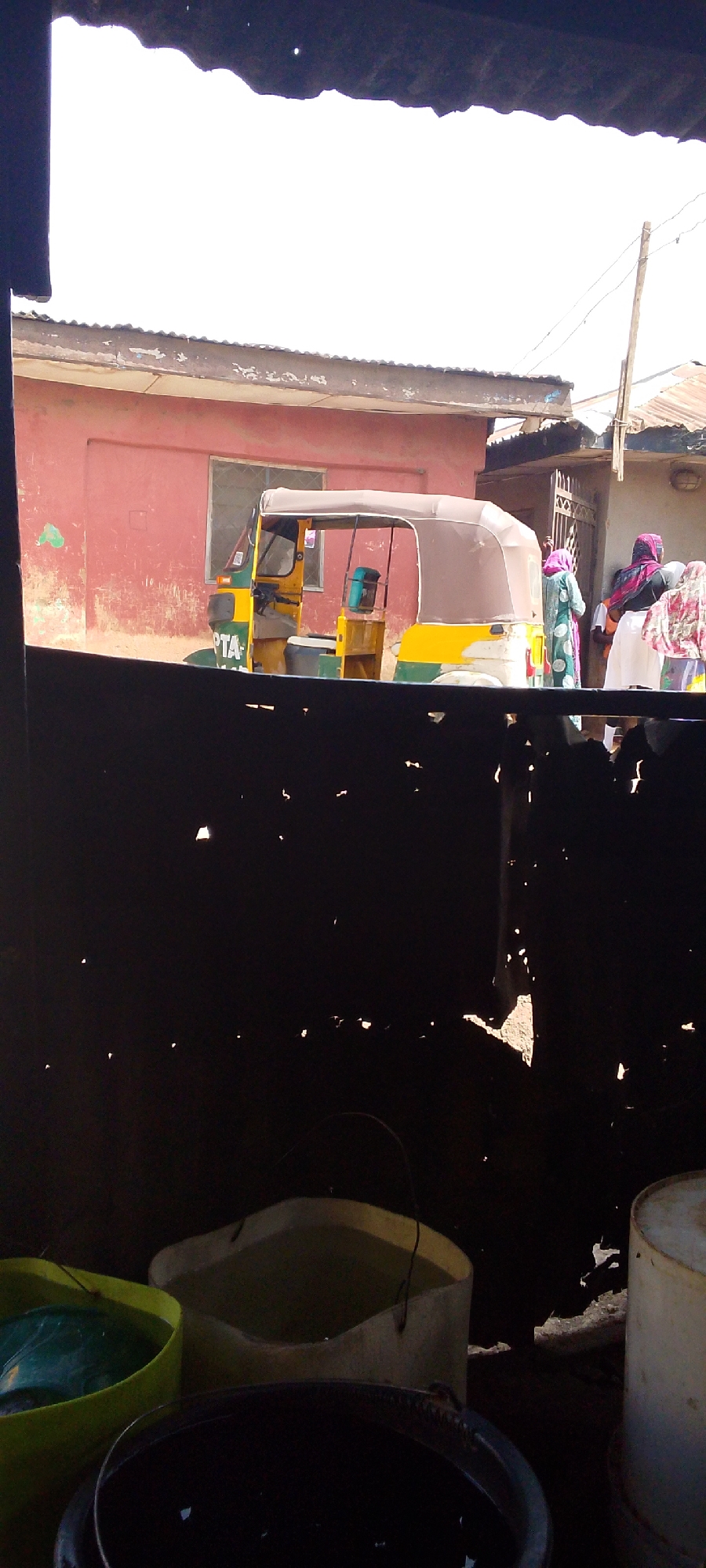 The image captures a vibrant street scene in what appears to be a residential or commercial area, viewed from behind a dark, perforated barrier. In the foreground, several plastic buckets or containers, some filled with liquid, are visible below a dark, possibly corrugated metal wall with numerous small holes allowing sunlight to penetrate.
In the midground, a yellow and green three-wheeled auto-rickshaw, commonly known as a Keke NAPEP in Nigeria, is parked. It features a light-colored canvas canopy. Partially visible text on the side of the vehicle reads "PTA". The auto-rickshaw is positioned in front of a reddish-brown, single-story building with a single window and a corrugated metal roof.
Further in the background, to the right, a small group of people, predominantly women, are gathered near the doorway of another building. They are dressed in long, flowing garments and head coverings, typical attire in parts of Nigeria. A wooden utility pole with overhead wires extends upwards on the right side of the frame.
The scene is illuminated by bright, natural daylight, indicating it is daytime. The clear lighting and visible sky suggest fair weather. The architecture, the presence of the auto-rickshaw, and the traditional clothing of the people are consistent with the provided location context of Jos, Nigeria, depicting a typical urban or semi-urban street. No specific activity or strong emotions are evident; it appears to be a moment of everyday life.