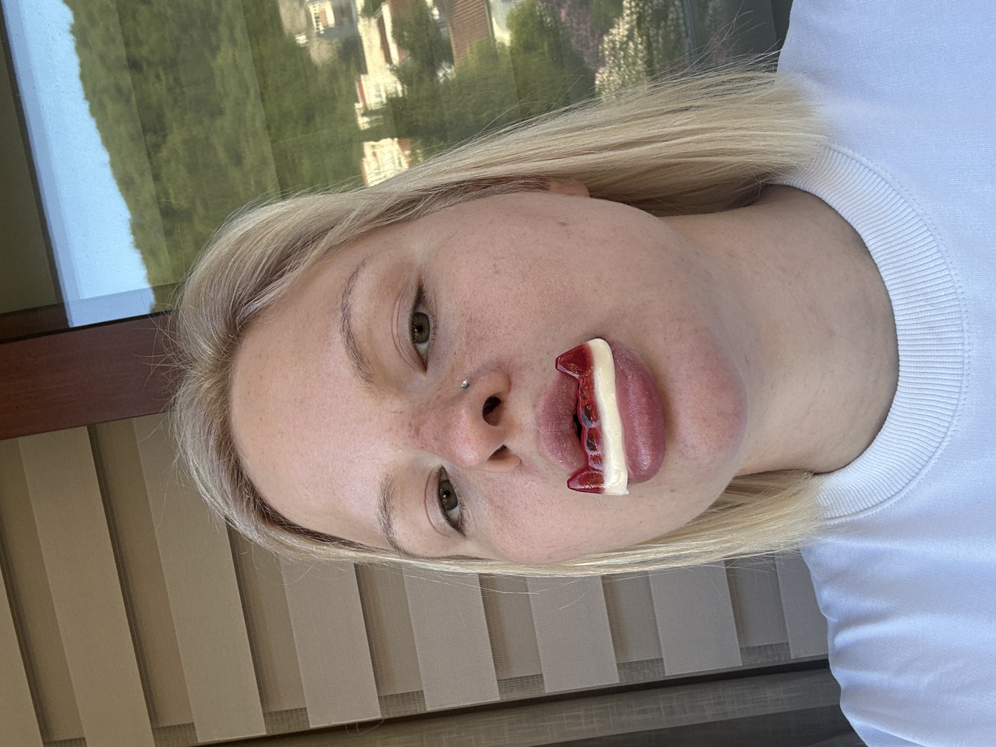 The image is presented vertically, featuring a young woman whose head is rotated 90 degrees counter-clockwise from an upright position. She has blonde, shoulder-length hair and fair skin with visible freckles. She wears a plain white crew-neck t-shirt. A silver stud nose piercing is visible. Her light brown eyes are open, looking towards the viewer. Playfully, she holds a red and white candy, resembling vampire fangs, in her mouth, partially visible between her lips.
The background behind her head features a large window on the upper left, revealing a bright outdoor scene with green trees and distant buildings under a sunny sky. Below her head, and to the right, are beige vertical blinds or shades. The overall lighting suggests daytime. The woman's expression is neutral, and there is no visible text. The setting appears to be indoors, near a window, with a view of a city or landscape, identified as Budva, Montenegro.
