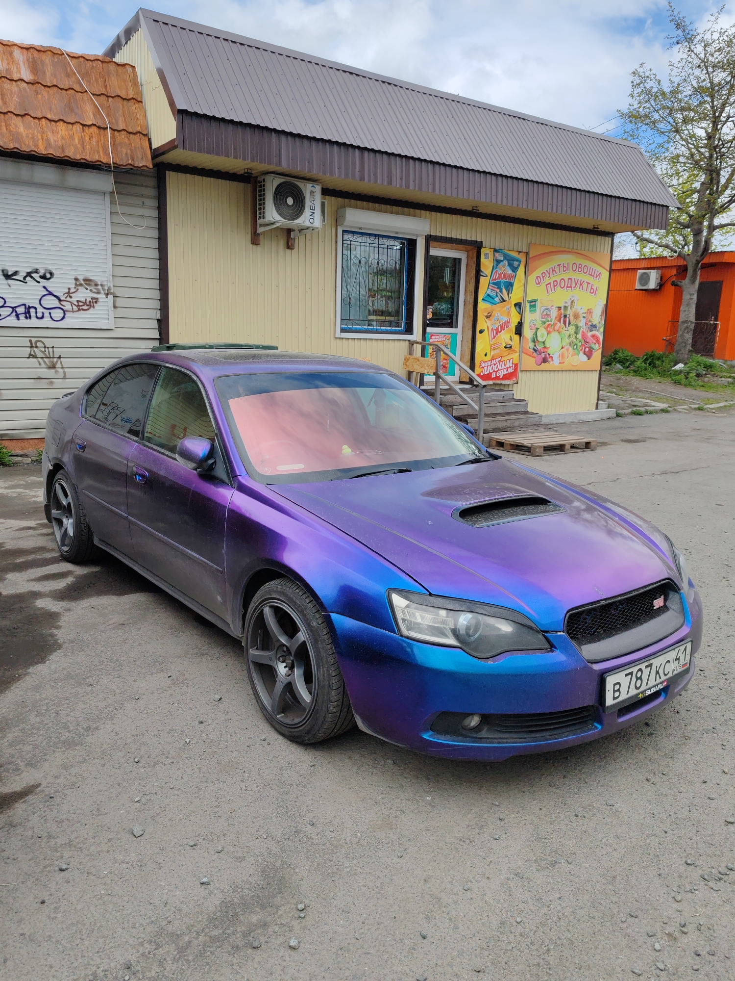 An eye-level, medium shot captures a purple and blue iridescent Subaru Legacy sedan parked on a rough, gravelly surface in front of a small, single-story commercial building. The building has a light yellow facade and a dark brown metal roof. To the left of the building, a window is covered by a white roller shutter adorned with graffiti. An air conditioning unit is mounted on the building's facade. To the right of the building's entrance, a large sign displays colorful images of fruits and vegetables with Russian text. A tree with green foliage stands to the far right. The car has a prominent hood scoop and its license plate reads "B 787 KC 41 RUS". The vehicle's interior is covered by a pink sunshade. The sky above is partly cloudy.