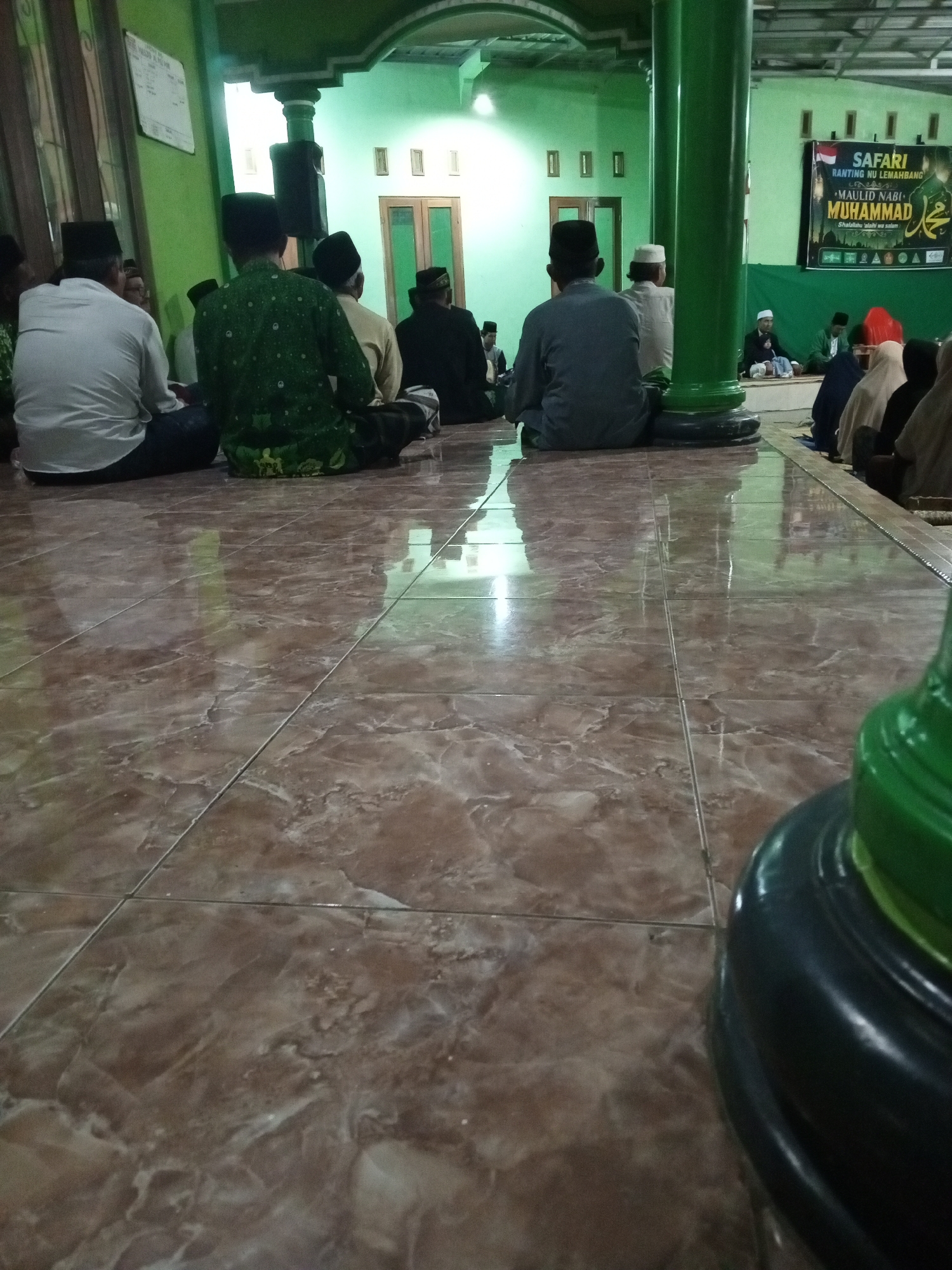 The image depicts a gathering of people, mostly men wearing traditional Muslim attire, including peci hats, seated on the floor inside a building that appears to be a mosque or Islamic center in Lemahbang, Indonesia. The room has green walls and a patterned tiled floor. In the background, a banner with text in Indonesian, including "SAFARI RANTING NU LEMAHBANG" and "MAULID NABI MUHAMMAD," is visible, indicating a religious event celebrating the birth of Prophet Muhammad. Several individuals are facing away from the camera, suggesting they are engaged in prayer or listening to a speaker. The overall atmosphere suggests a communal religious observance.