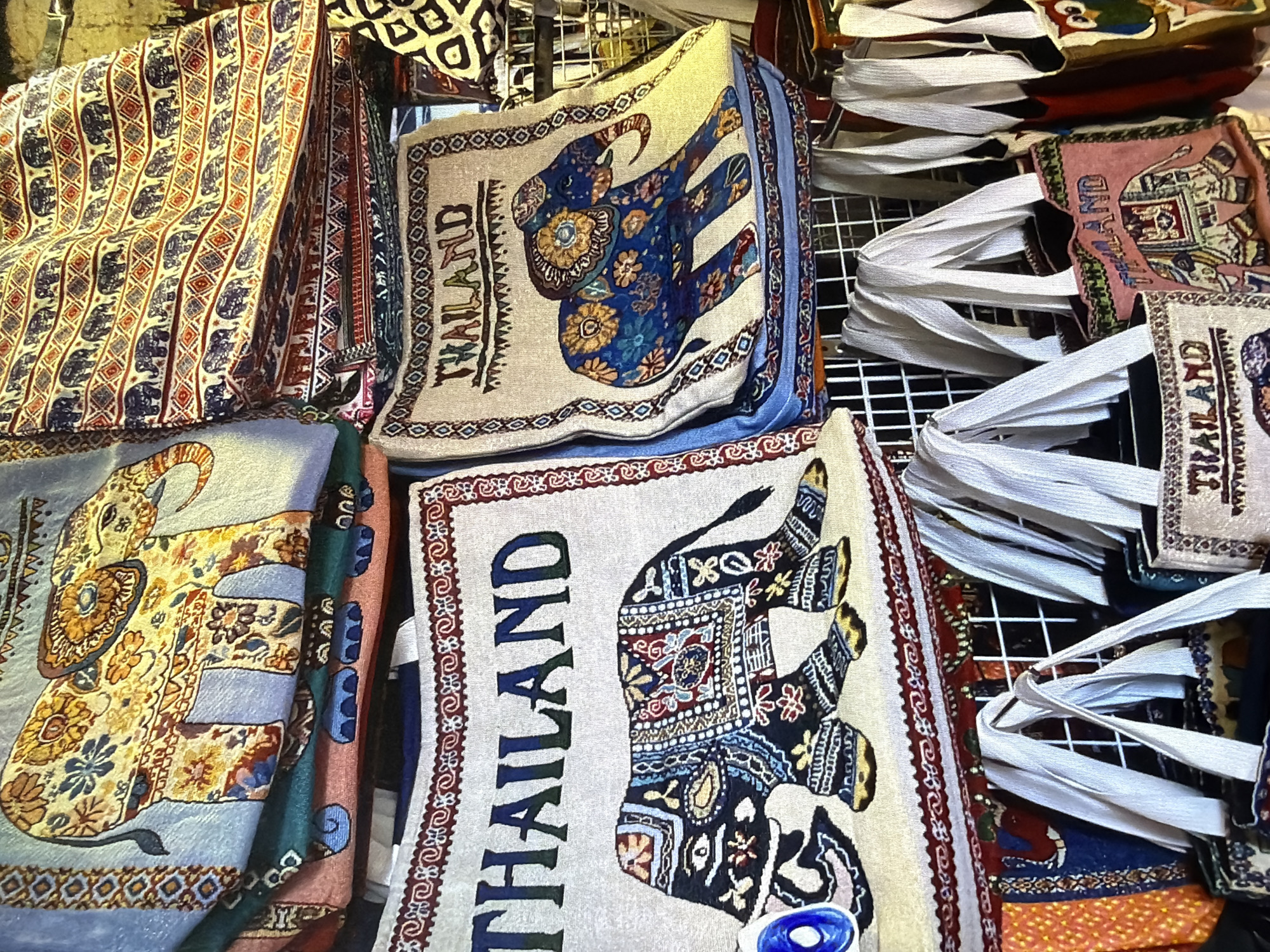 This image captures a close-up view of numerous fabric items, likely bags or textiles, arranged for sale or display. The predominant theme across many of the items is an elaborately decorated elephant design. These elephants feature intricate patterns, often in floral or geometric motifs, and are rendered in various colors such as blue, gold, black, and red against backgrounds of beige, light blue, or pink.
Several of the items clearly display the word "THAILAND" in a bold, stylized font, running vertically along one side of the fabric. This indicates their origin or theme as souvenirs from Thailand. The fabrics themselves appear to be made of a woven material, possibly tapestry-like, giving them a textured appearance.
The items are stacked and folded, showing different sides and patterns. In the upper left, a pile of textiles features a repeating pattern of smaller elephants and geometric lines in red, blue, and cream. Towards the right, a vertical stack of white fabric handles suggests that many of these items are tote bags or similar carriers. A metal grid or rack is partially visible in the background, reinforcing the idea of a market stall or shop display. The lighting appears to be indoor or artificial, highlighting the vibrant colors and detailed designs of the textiles.