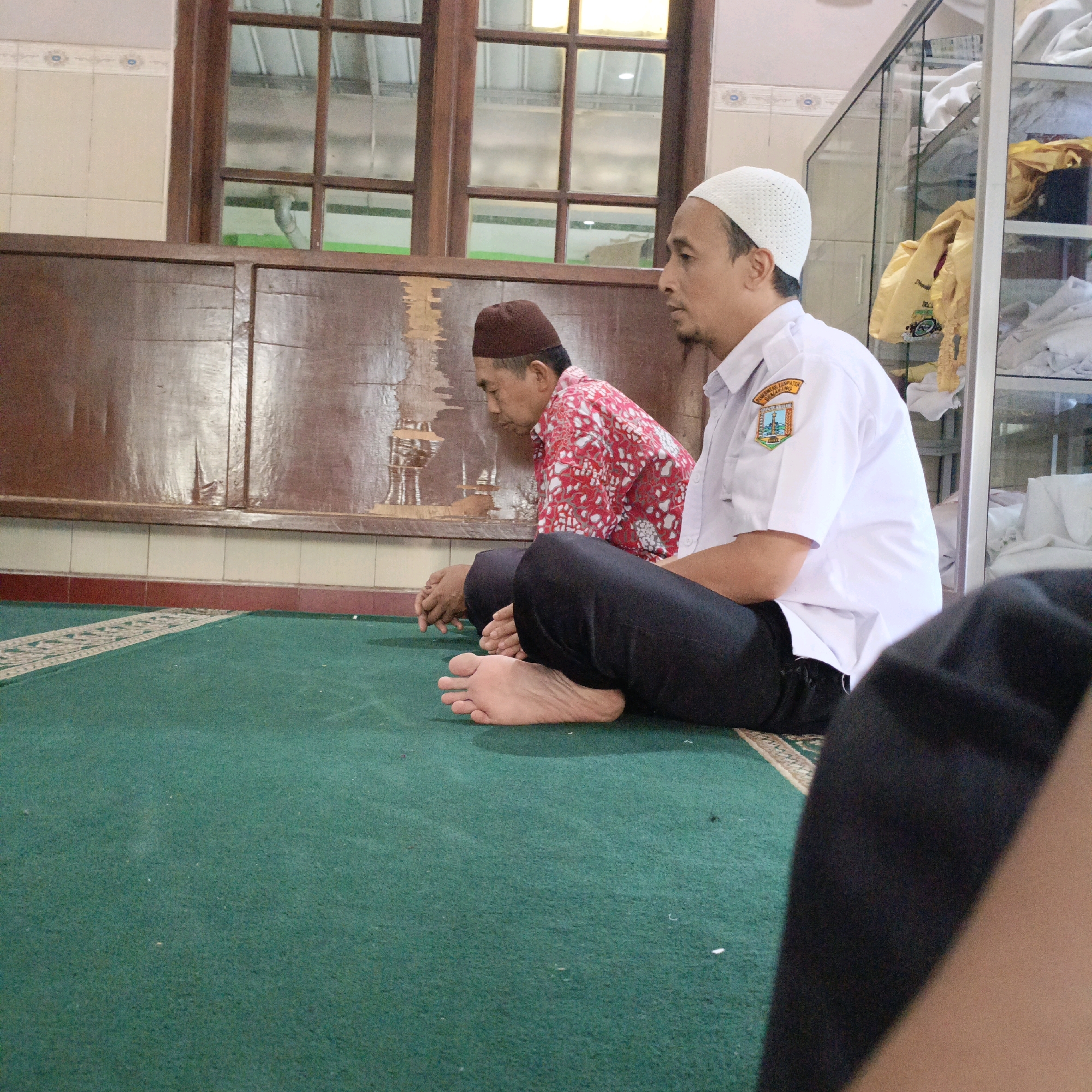 In an indoor prayer area, two men are seated on a green carpet, facing away from the camera. The man on the right wears a white skullcap and a white collared shirt with a crest on the left breast, reading "PEMERINTAH KABUPATEN SEMARANG" and depicting a local emblem. He is wearing dark pants. The man on the left wears a brown skullcap and a red shirt with a white pattern. He is also wearing dark pants. Both men are in a prayer posture with their hands resting on their laps. The background consists of a wooden wall with a horizontal wooden beam and a series of large wooden-framed windows. To the right of the men, a glass display cabinet is visible, containing folded garments, some of which are yellow. The overall impression is of a simple, functional prayer space. The presence of the "PEMERINTAH KABUPATEN SEMARANG" crest on the shirt suggests the location may be within the administrative boundaries of Semarang Regency, which is adjacent to Salatiga, Indonesia. However, the exact location within Salatiga cannot be definitively determined from the image alone.
