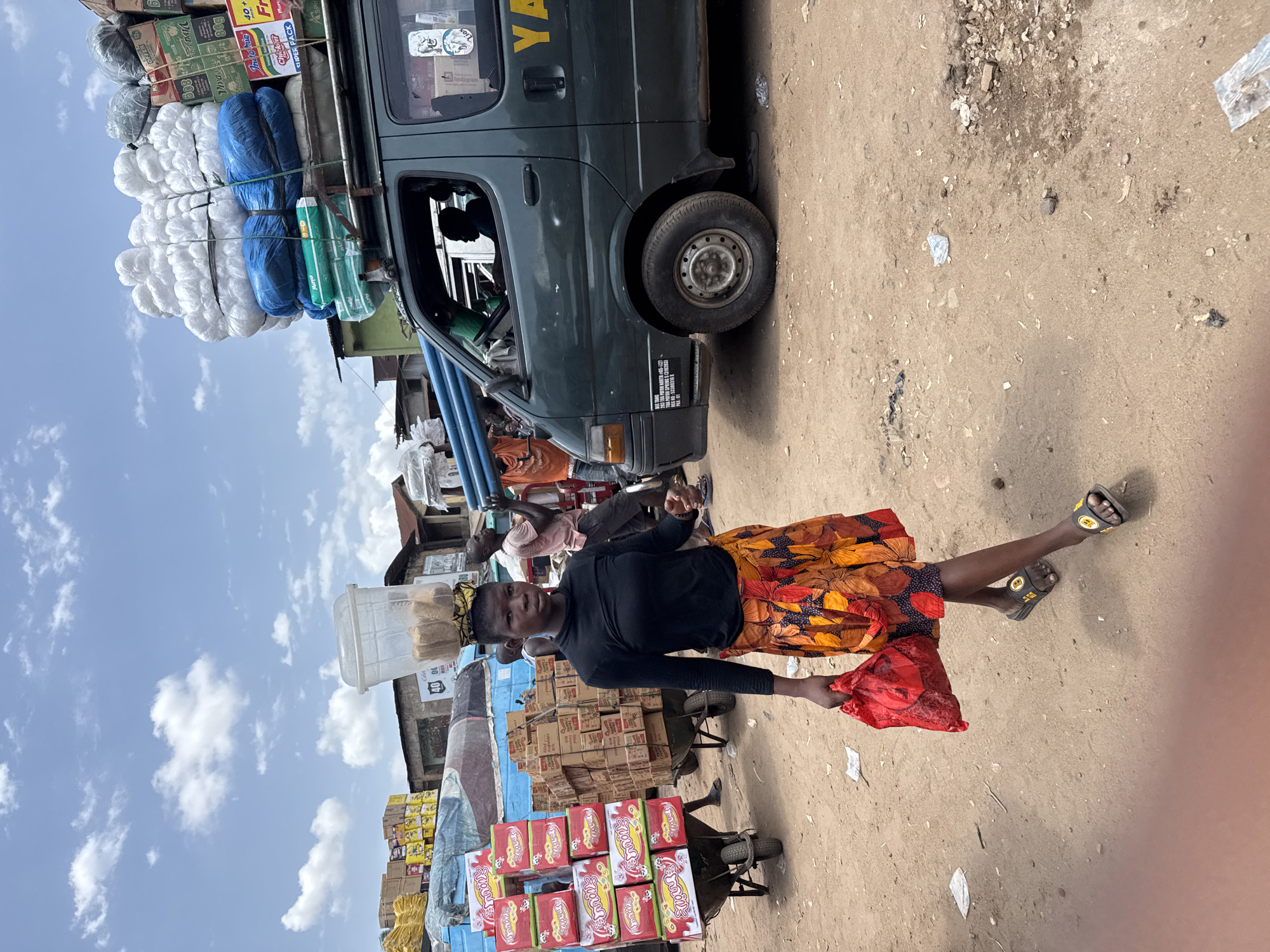 The image shows a woman pulling a cart laden with merchandise in what appears to be a bustling street market in Onitsha, Nigeria. The cart is stacked high with boxes and other items, including boxes of "Indomie" noodles. The woman is wearing a long-sleeved black shirt and an orange-patterned skirt. She is holding a red plastic bag in her left hand. On her head, she is carrying a white plastic container with brown content.
Behind her is a dark green van with goods piled on its roof, including bundles of what looks like white yarn wrapped in blue plastic. Other people are visible in the background, suggesting a busy commercial area. The ground is unpaved and dusty, with scattered debris. The sky is partly cloudy.