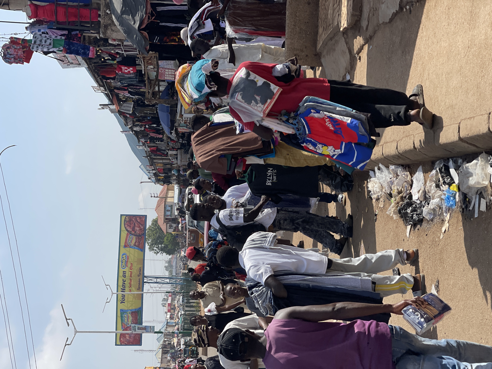The image depicts a crowded street scene in Bukuru, Nigeria. A line of people stand along the sidewalk, most carrying bundles of clothing on their shoulders. Many appear to be hawkers or vendors displaying their wares. The clothing is diverse, including both casual wear and more formal attire. A large advertising billboard for "ONGA" food products is visible in the background. There is trash visible on the ground near the sidewalk. In the background there are buildings of varying heights. The sky is partly obscured by the buildings and the stalls on the left.