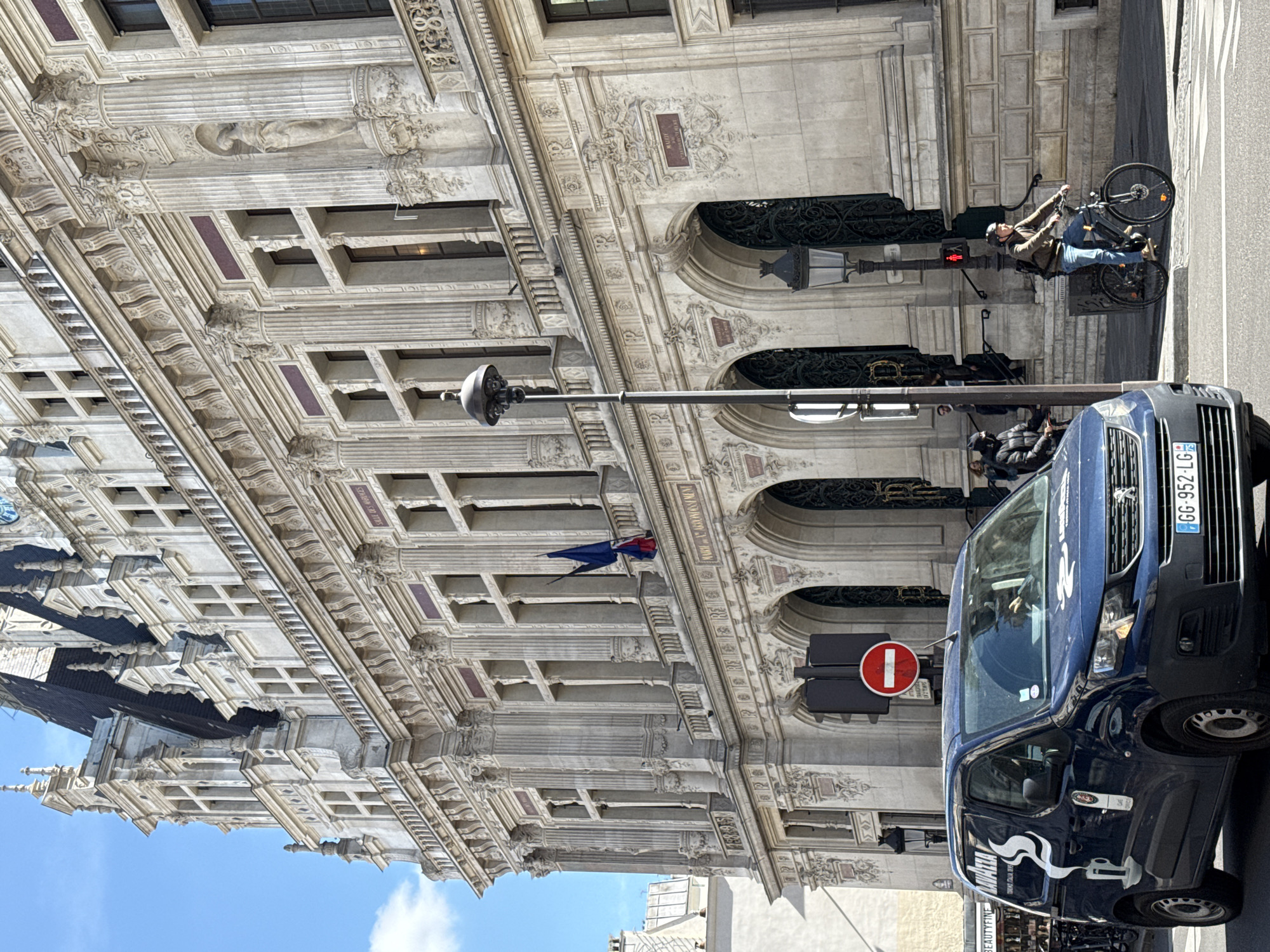 The image depicts a street scene in Paris, France. An ornate, multi-story building with elaborate architectural details is the backdrop. A blue Peugeot van is parked on the right side of the frame. A man on a bicycle is present in the background. A "no entry" traffic sign is visible near the van, and a flag is hanging from the building.