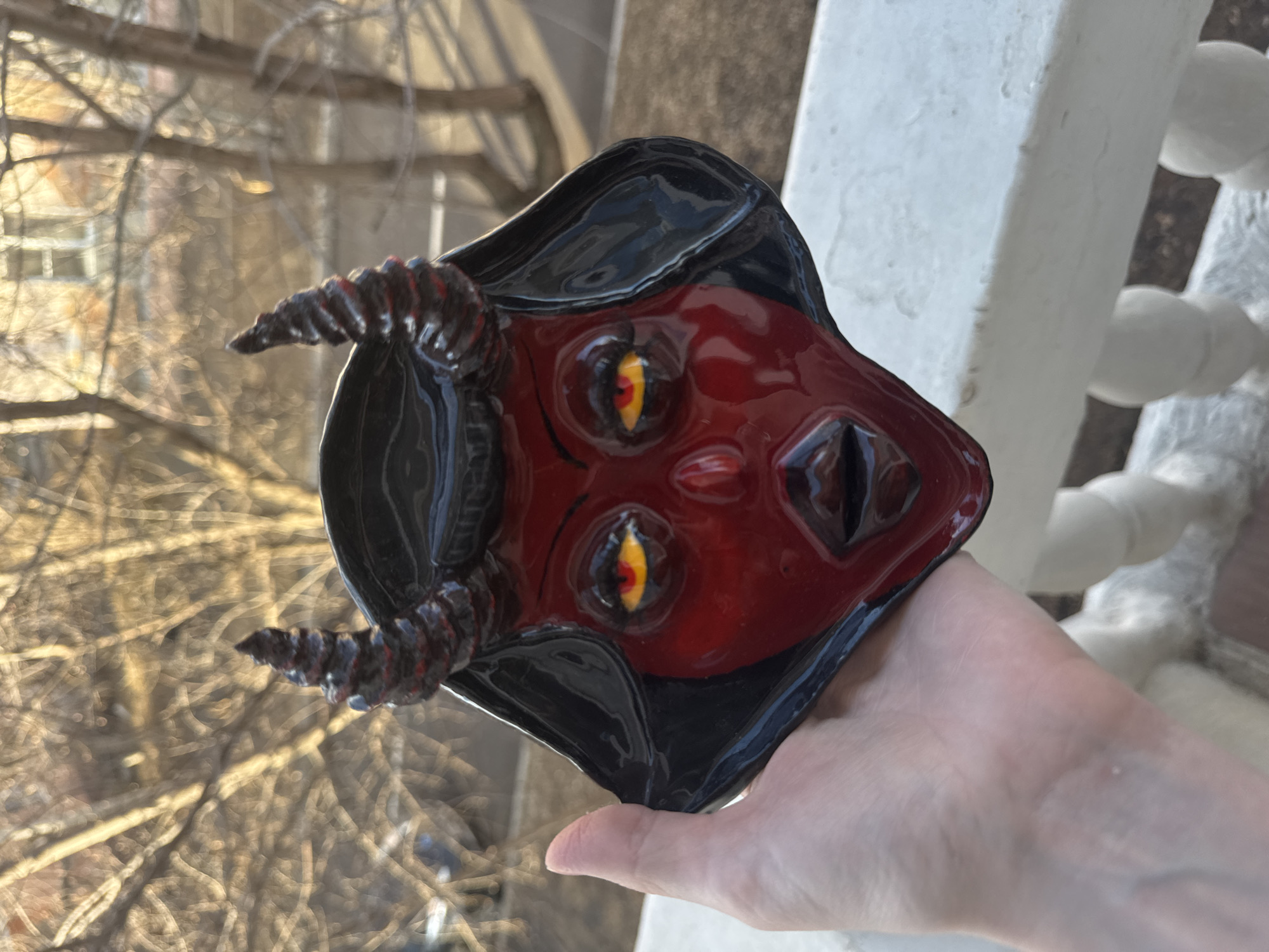 A hand holds a glossy, glazed ceramic mask resembling a devilish face with horns and a black hood. The mask has red skin, yellow eyes, and black accents. The background shows a blurred outdoor scene of leafless trees and a white-painted balustrade. There is no information to suggest it is taken in Krasnoyarsk, Russia.