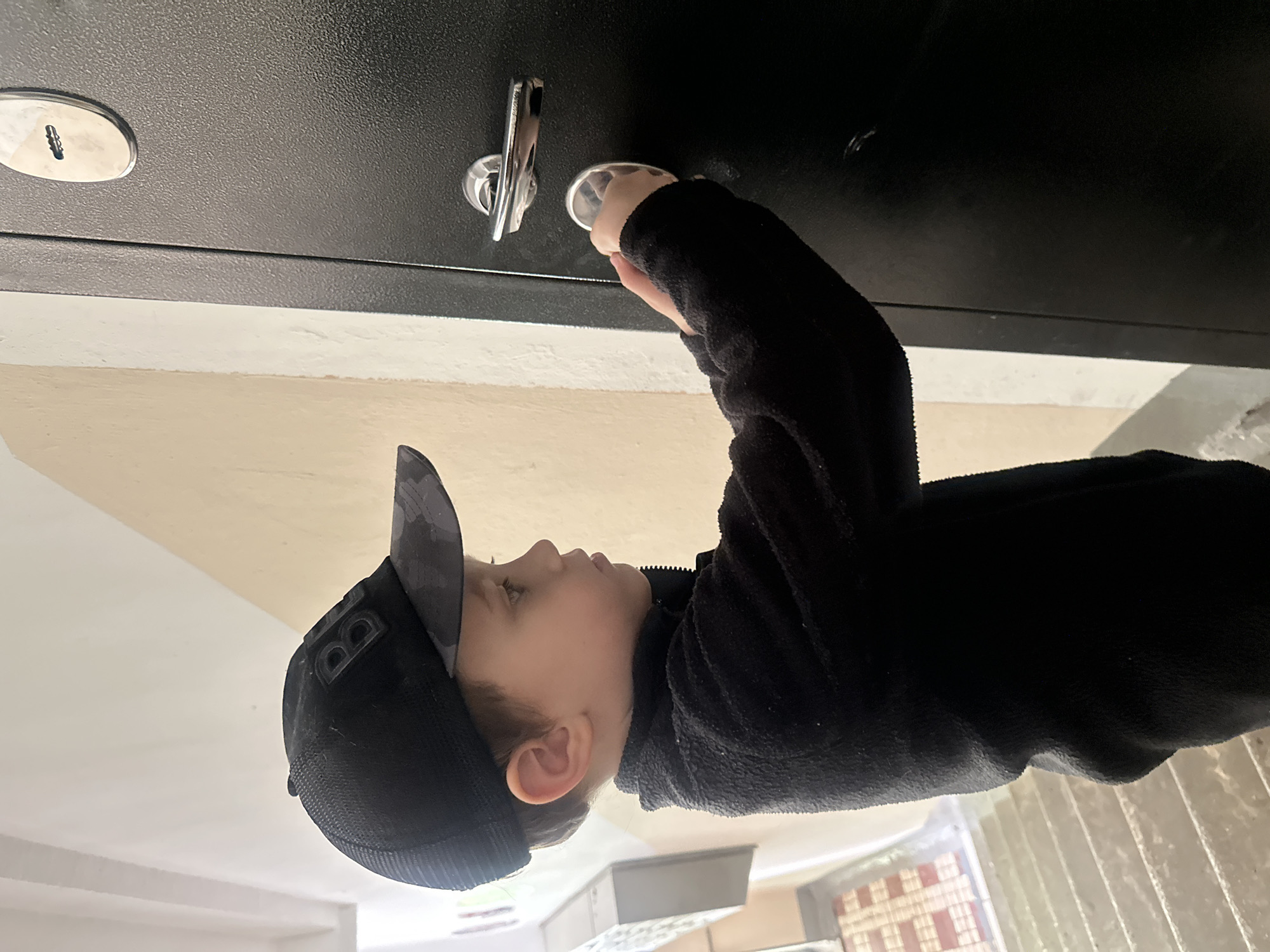 A young boy wearing a black fleece jacket and a black baseball cap with a camouflage brim is reaching for the handle of a dark metal safe. The safe has two keyholes and a chrome handle. The boy's face is partially visible, and he appears focused on the task. The image is shot from a slightly low angle, with the safe positioned above the boy. The background shows a light-colored wall and the top of some stairs. The lighting suggests it is daytime, possibly indoors or in a shaded outdoor area. The overall scene conveys a sense of curiosity and exploration. There is no visible text in the image.