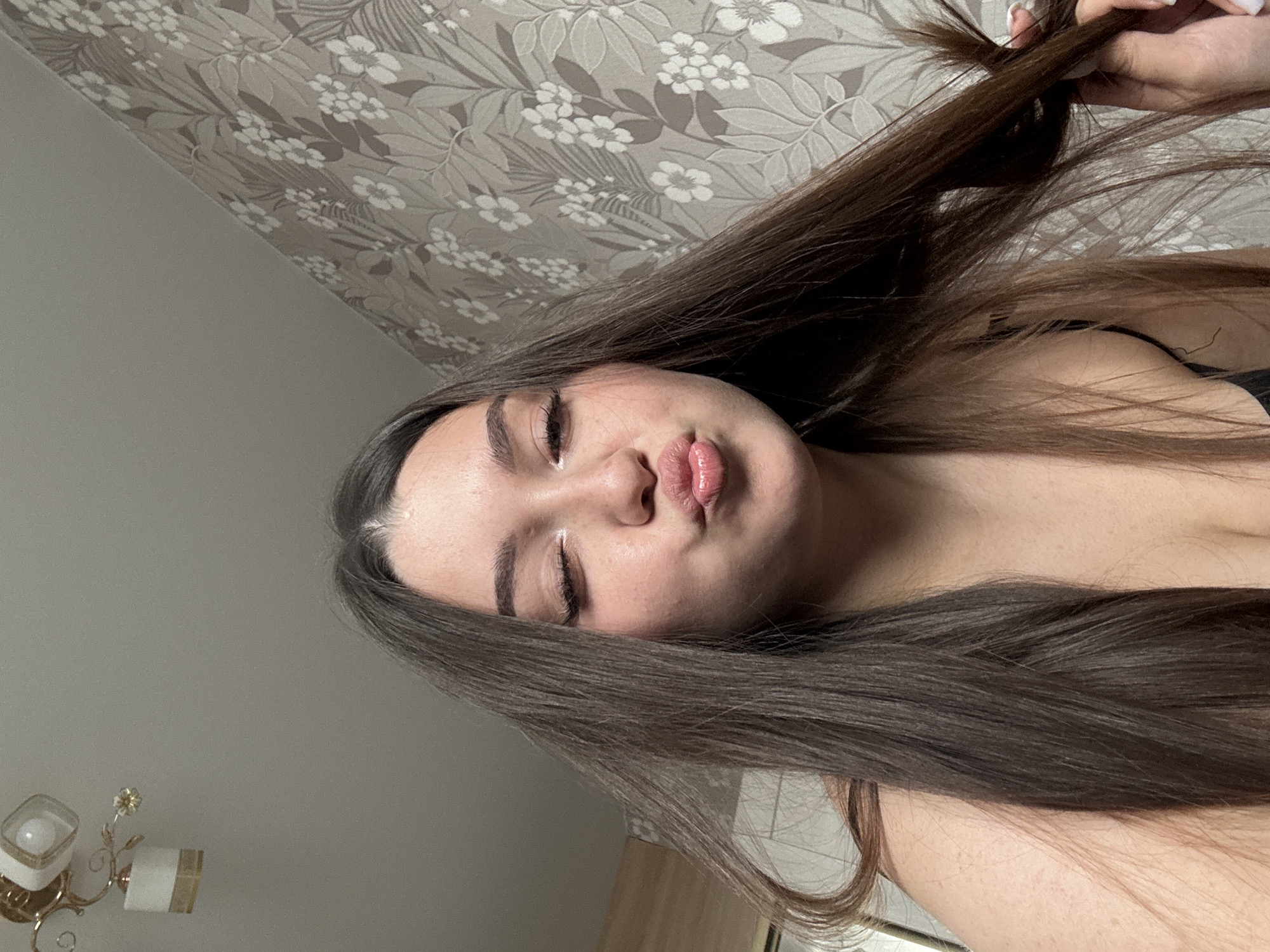 A low-angle, color, indoor portrait photograph shows a young, light-skinned woman with long, brown hair. The woman's hair is parted slightly to the side and she is holding some of it in her right hand, near her hair ends. She has closed eyes and pouty lips. Light appears to be reflected on her forehead and lips. The background is a gray and white floral wallpaper. To the lower left, a gold and white wall sconce is visible. A gray wall is to the lower left. The photograph appears to be taken indoors in a room, and no specific location in Perm, Russia, can be determined from the photograph.