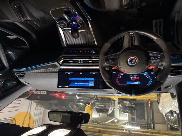 The image presents the interior of a modern performance vehicle, specifically from the driver's perspective, inside a brightly lit underground parking garage in Warsaw, Poland.
The foreground is dominated by the vehicle's cockpit. The multi-function steering wheel features a black leather rim with alcantara/suede grips, carbon fiber accents, and a distinctive Alpina emblem at its center. A blue stripe marks the 12 o'clock position, and red paddle shifters are visible behind the wheel. The digital instrument cluster displays vehicle status, including a warning for an open door and a graphical representation of the car. To the right, the infotainment screen shows "Добро пожаловать, гость!" (Welcome, guest!) in Russian, along with the time (19:07) and signal indicators. The dashboard features carbon fiber trim and a continuous strip of blue ambient lighting. The center console includes a modern gear selector, an iDrive controller, and an engine start/stop button, all surrounded by carbon fiber materials. No occupants are visible within the vehicle.
Beyond the windshield, the underground parking garage is illuminated by artificial fluorescent lighting. The concrete ceiling shows exposed pipes and ventilation systems. The concrete floor is marked with yellow lines indicating parking spaces. Several vehicles are parked in the background: a white SUV on the left, a dark sedan further back, a grey SUV to its right, and a black BMW sedan positioned to the right with a visible Polish license plate "WH 2697F," confirming the location in Warsaw. A large, abstract yellow and orange graphic, resembling cutlery (a fork and knife), is painted on a concrete wall in the background to the right. Red and white striped bollards are also visible near the white SUV.