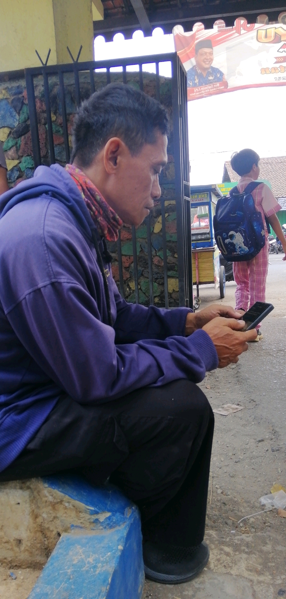 In Ajibarang, Indonesia, a man wearing a purple hoodie and black pants is sitting on a blue concrete structure and looking at his phone. He has short, dark hair and is wearing a patterned scarf around his neck. In the background, a young child in a pink striped outfit with a backpack is walking away from the camera. There's a food cart and a fence with colorful stones visible. The scene appears to be outdoors, with a light-colored building and a banner in the distance.