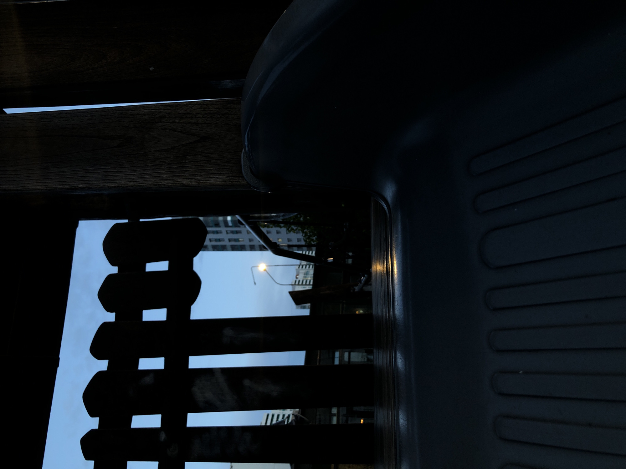This media file captures an urban scene at dusk or night, viewed from an obstructed perspective. The image is predominantly dark, with strong contrasts between silhouetted foreground elements and the slightly brighter background.
In the bottom right, a dark, smooth object, possibly a plastic or metal chair backrest or seat, fills a significant portion of the frame. It exhibits subtle ridges or grooves on its surface, indicating its texture and shape. To the left, a dark wooden slatted structure, resembling a fence, screen, or part of a bench, stretches horizontally across the mid-ground. The wood grain is faintly visible, but the structure is largely a silhouette.
Beyond these foreground elements, an urban landscape unfolds. The sky, visible through and above the slats, is a light blue, suggesting the lingering light of dusk or early morning. Tall, grey buildings with numerous windows, characteristic of a city, rise into the sky. One prominent, illuminated streetlight with a yellow glow stands out against the backdrop, reinforcing the impression of evening. Some dark, undefined shapes, likely trees or bushes, can also be discerned amidst the urban structures.
The image does not depict any specific activity or event, but rather offers a quiet, observational view of a city winding down for the night. The time of day appears to be dusk or night, given the active streetlight and overall low light. The weather is clear, with no clouds visible. The location cues point to an urban setting, consistent with Saint Petersburg, Russia, as provided in the context. There is no visible text in the image.