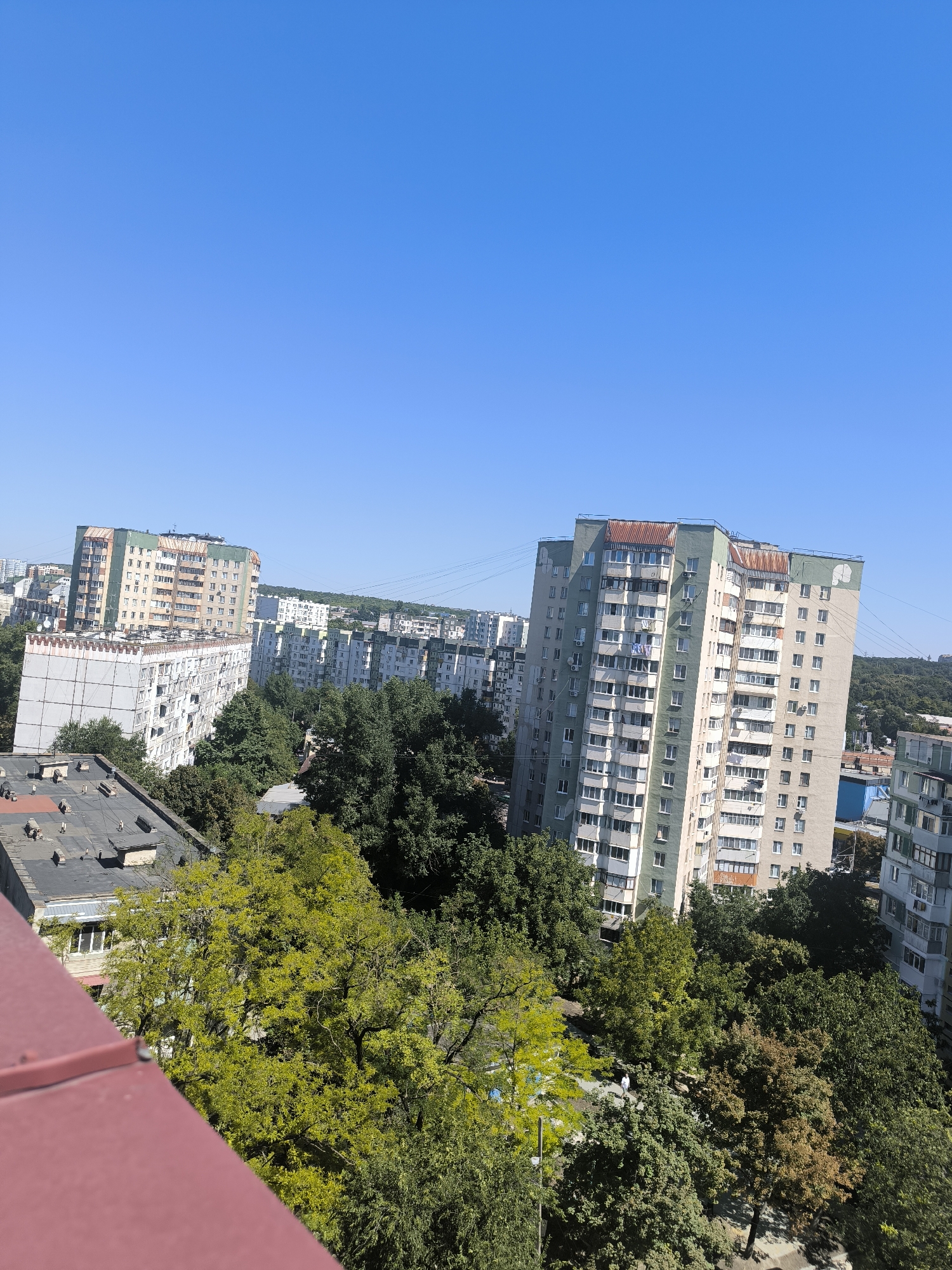 This is an aerial, eye-level view of a residential area in Chișinău, Moldova, under a clear blue sky. In the foreground, a red-tiled roof occupies the bottom left corner. Behind it, a multi-story apartment building with a flat, dark roof, featuring ventilation shafts and what appear to be satellite dishes, is visible.
Further into the scene, several more tall apartment buildings dominate the landscape. One prominent building in the mid-ground is light green and beige with many balconies and windows. To its left, a lighter colored, rectangular apartment building with a patterned facade is visible. Behind these, a series of identical, light-colored apartment blocks stretch across the horizon, interspersed with dense green trees. The trees vary in color, with some exhibiting bright green foliage and others a more muted, autumnal hue. A single person, dressed in white, is walking on a path amidst the trees in the lower right portion of the image. The presence of various apartment buildings, the types of trees, and the general architectural style are consistent with residential neighborhoods found in Chișinău.