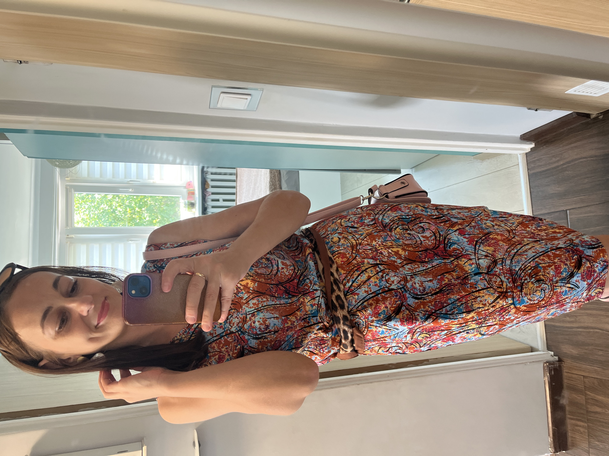 The image shows a woman taking a selfie in what appears to be a home setting. She is wearing a vibrantly colored, patterned dress with a leopard print belt. A light pink purse is slung over her shoulder. The background suggests an indoor environment with natural light coming from a window. The woman is smiling and touching her hair, appearing relaxed and happy. The image is oriented vertically. There is no visible text in the image.