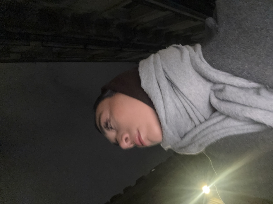 A woman wearing a dark head covering and gray shawl or sweater is partially visible. She is outside at night with a light source illuminating the bottom right of the frame. The sky is dark, and the image appears to be taken at an angle. The woman's face is in profile, and her expression is neutral. A dark, textured surface is visible at the top of the frame.