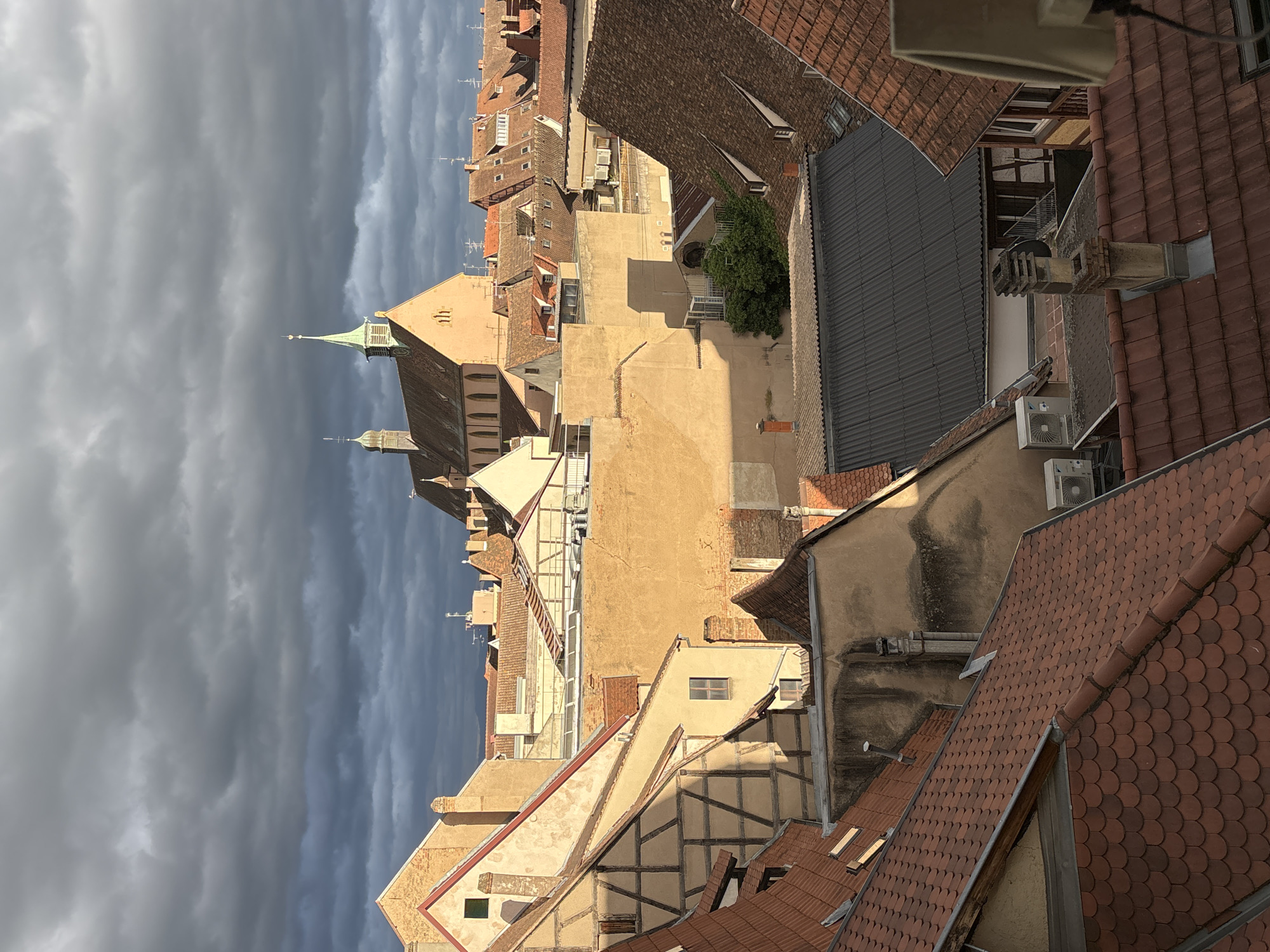 The image depicts a rooftop view of the historic city of Colmar, France, with a dramatic sky overhead. The architecture suggests the traditional half-timbered style common in Alsace, with steeply pitched terracotta tiled roofs. In the background, a church with a green steeple stands prominently, a common landmark in European cities. The lighting indicates it is either early morning or late afternoon, with strong shadows cast across the buildings. The presence of air conditioning units on some rooftops suggests modern additions to the older structures. The scene is devoid of people, focusing solely on the urban landscape and the interplay of light and shadow under a cloudy sky.