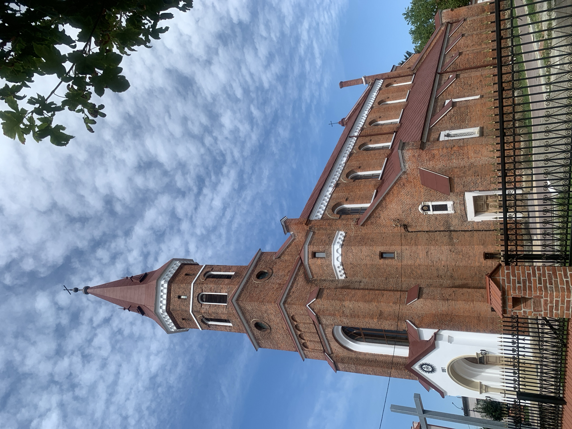 The image shows a red brick church with a tall, octagonal tower topped by a red, pointed roof and a cross. The church has arched windows and a white, arched entrance with the year "1897" inscribed above it. A large gray cross stands to the left of the entrance. The church is surrounded by a black, ornate metal fence. The sky is blue with scattered white clouds, and a tree branch with green leaves is visible in the upper right corner. The building is in the city of Kolomyia, Ukraine.