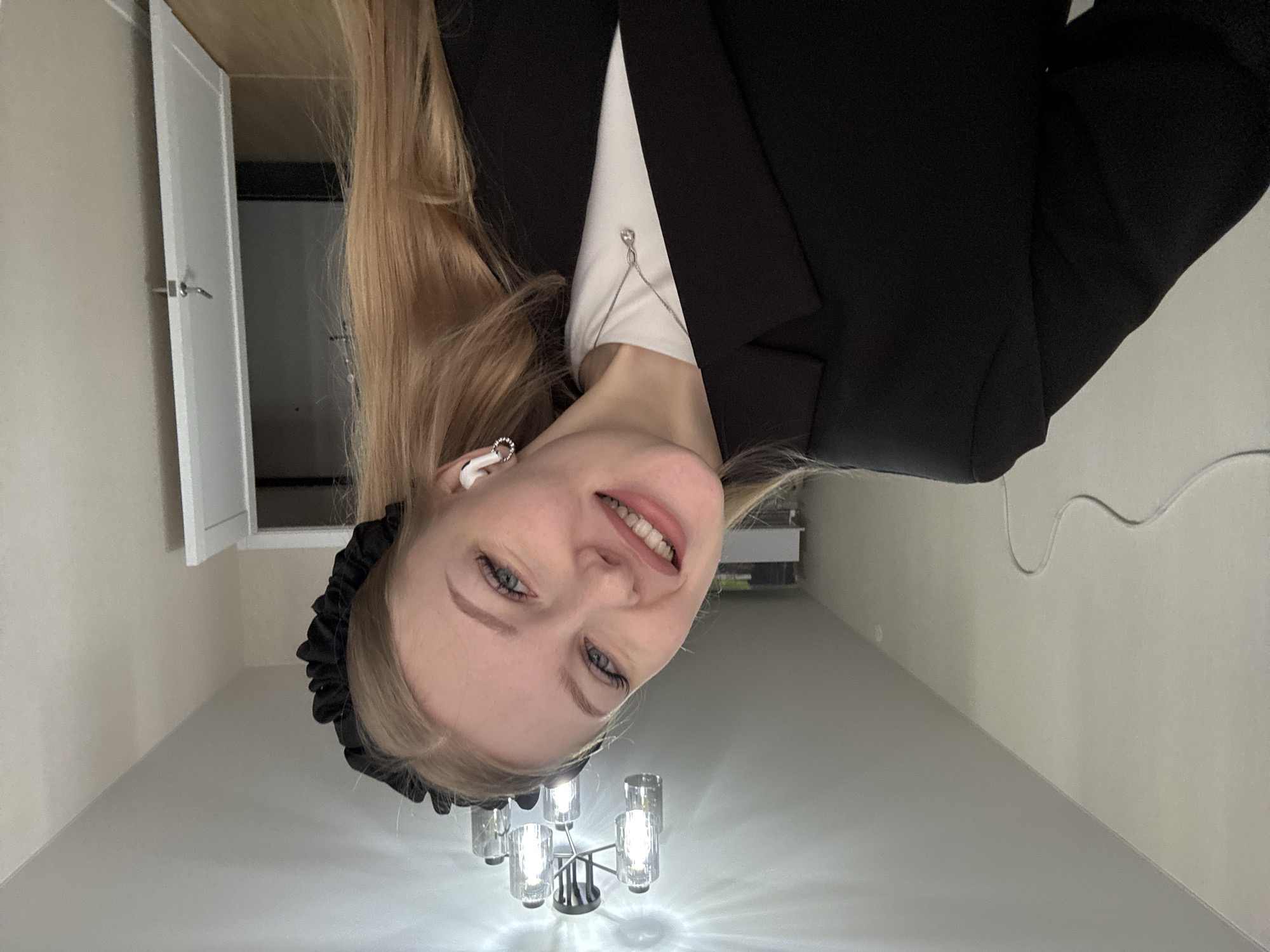 A blonde woman wearing a black blazer and a black ruffled headband is looking directly at the camera with a slight smile. She appears to be indoors under a chandelier with six frosted glass shades. She is wearing an earbud. In the background, there is a closed white door. There is an electrical cord visible along the wall. There is no evidence to determine if the image was taken in Perm, Russia.