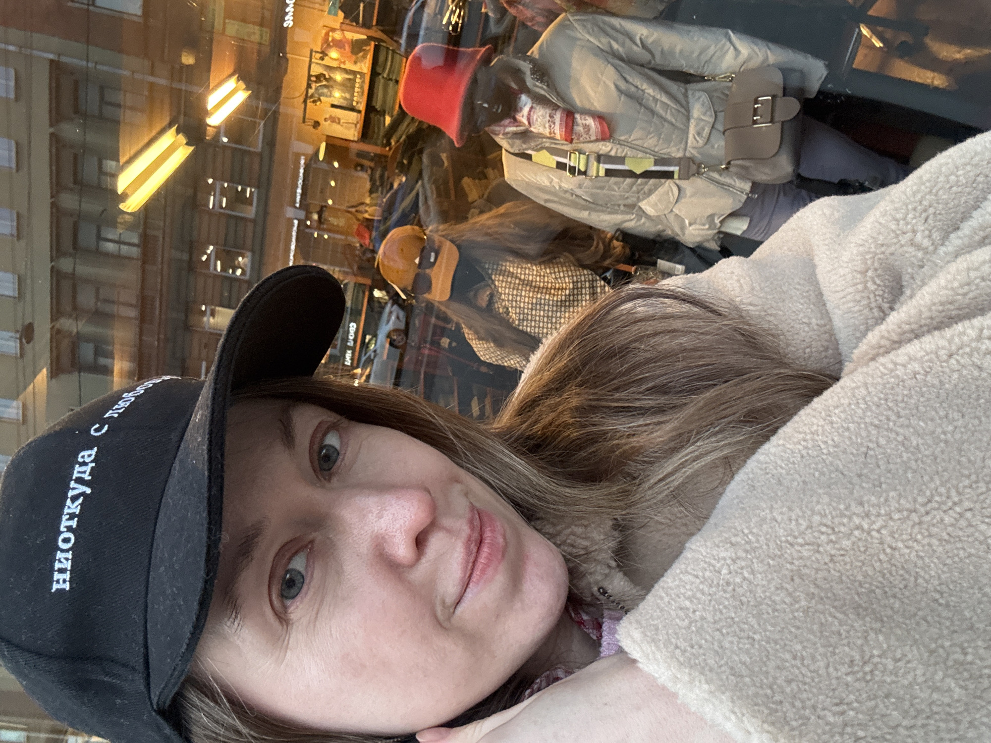 A woman is taking a selfie in front of a storefront window in Saint Petersburg, Russia. The window reflects buildings and streetlights. Behind the woman, mannequins are dressed in hats, jackets, and accessories in the store display. The woman wears a black baseball cap with white Cyrillic lettering and a beige fleece jacket.