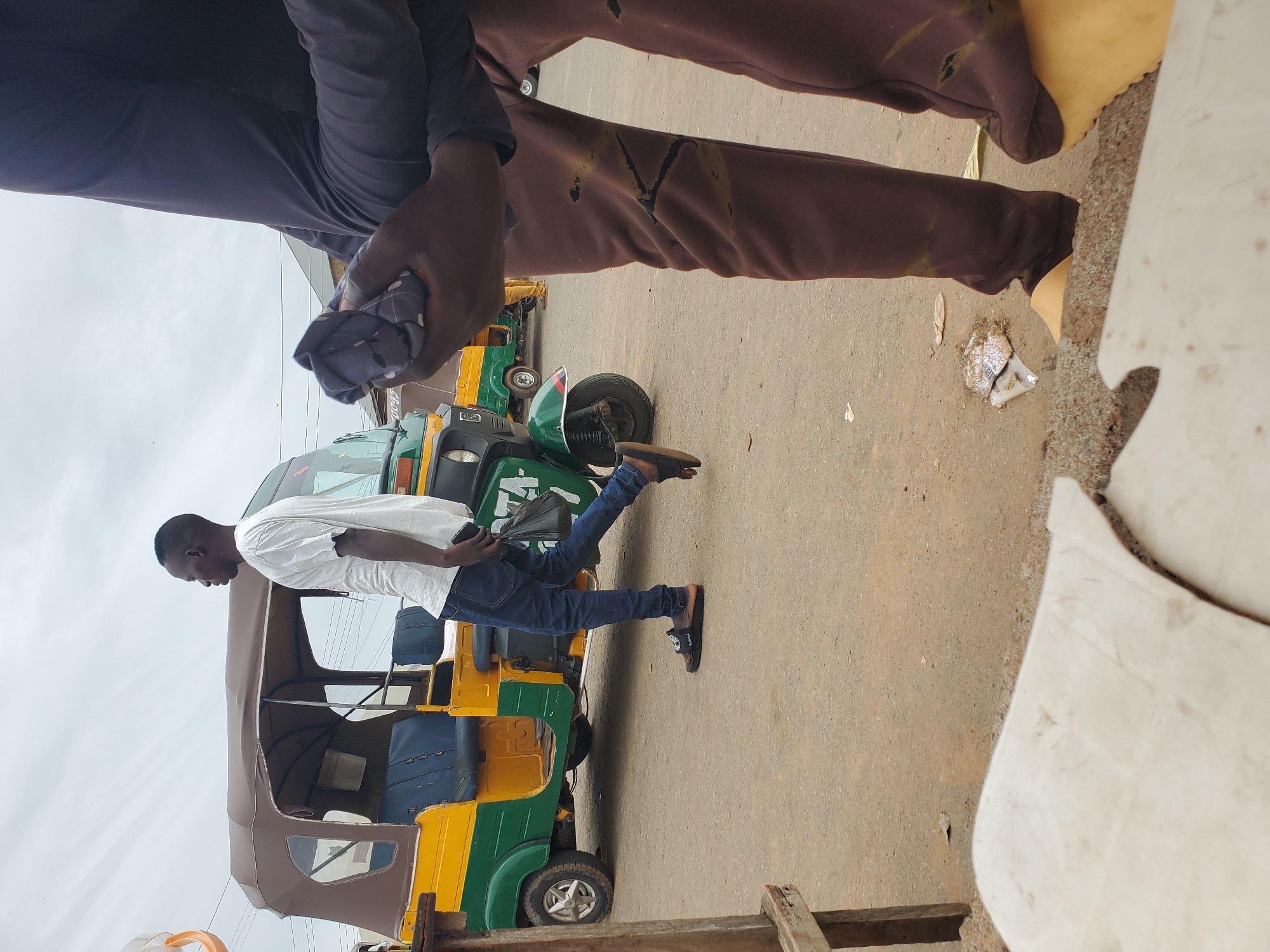 An outdoor scene in Gwong, Nigeria, captures a street view with a yellow and green auto-rickshaw parked on the right. A man in a white t-shirt and blue jeans is walking past the auto-rickshaw, carrying a black plastic bag in his right hand. He is wearing black sandals with brown straps. In the foreground, another person's lower body is visible, wearing brown sweatpants with dark markings and yellow footwear. The ground is paved with a rough, dirt-like surface, and scattered debris is present. The sky above is overcast with grey clouds.