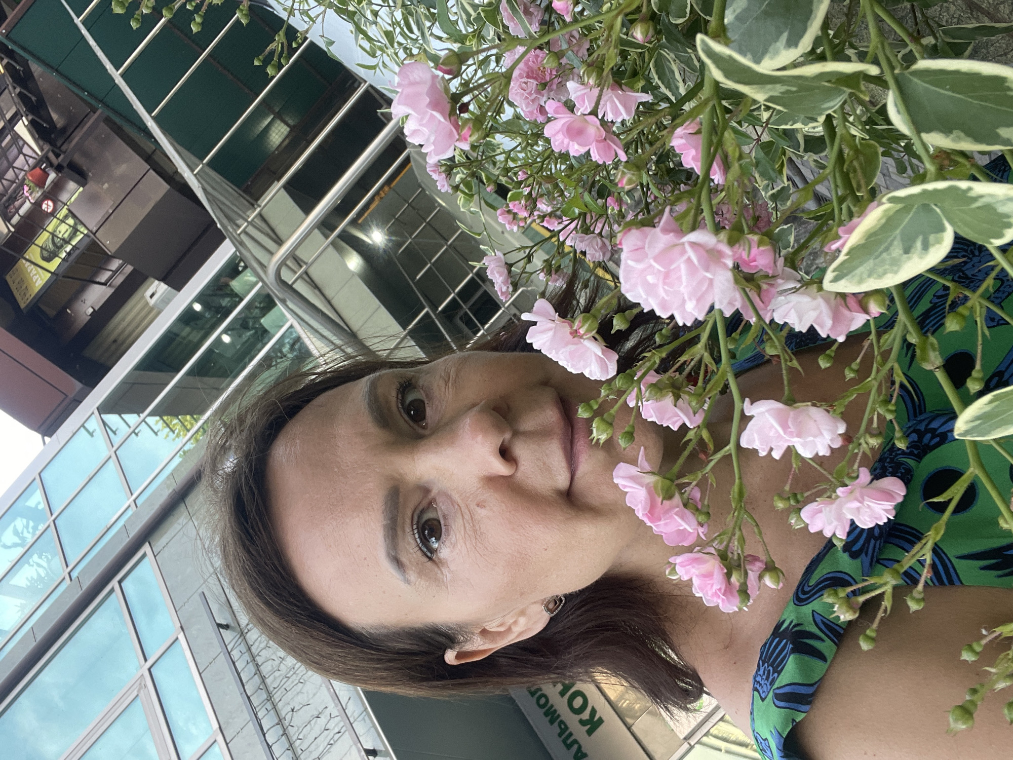 A woman is taking a selfie outdoors, holding a bouquet of pink roses. She has brown hair and is wearing a green and blue patterned dress. Behind her, a modern building with glass windows and balconies is visible, with a sign in Russian that reads "АЛЬМОГ КОН" which likely refers to a business or organization. The scene appears to be in a city, possibly Sochi, Russia, given the Russian signage and architectural style.