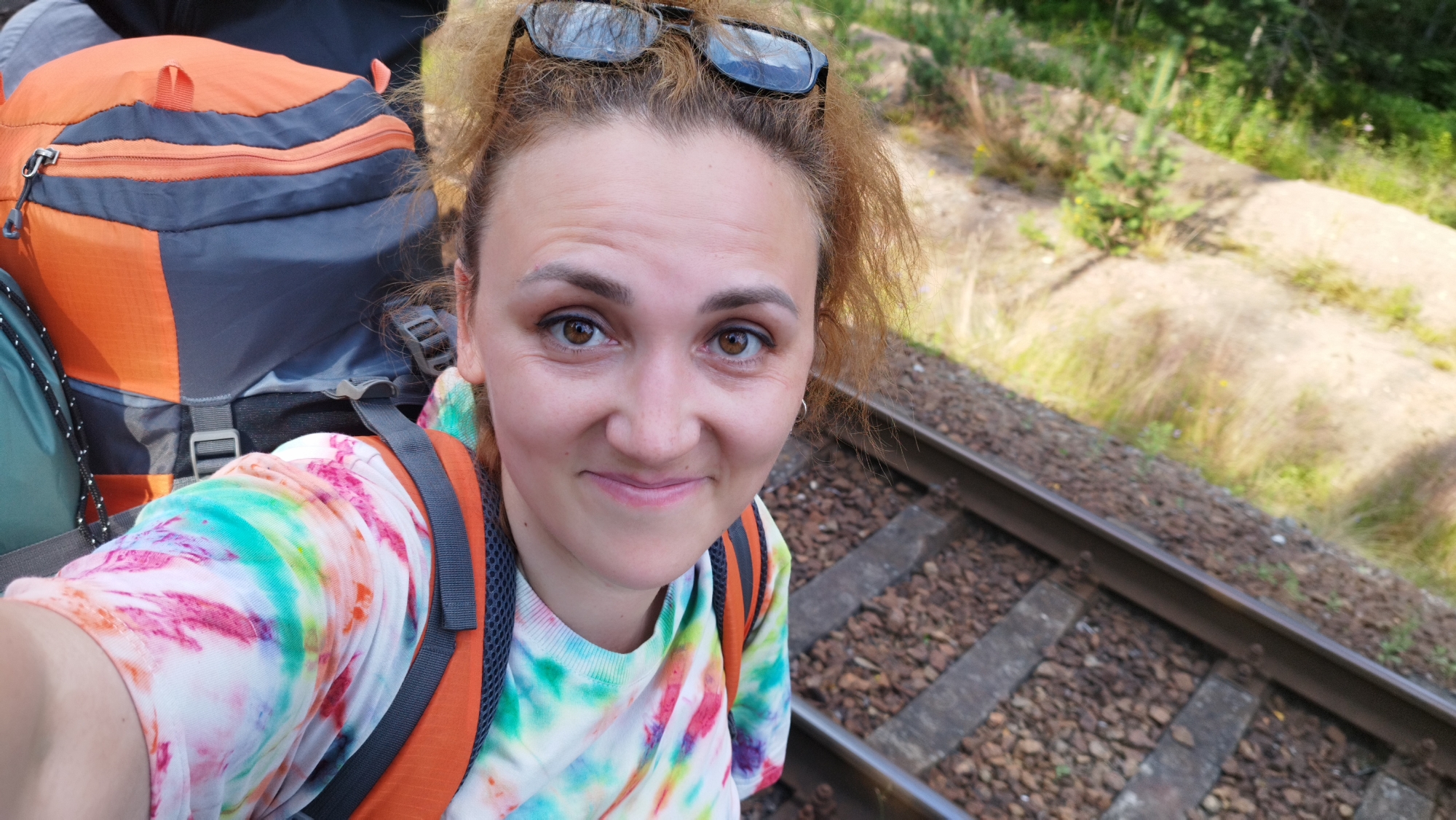 A woman with auburn hair and sunglasses perched on her head is taking a selfie while standing next to train tracks. She is wearing a tie-dye t-shirt with a backpack. The backpack is orange and grey. The train tracks are made of ballast and wood ties. The environment is outdoors with greenery and trees visible in the background. The location is the city of Tarasovskoe, Russia.