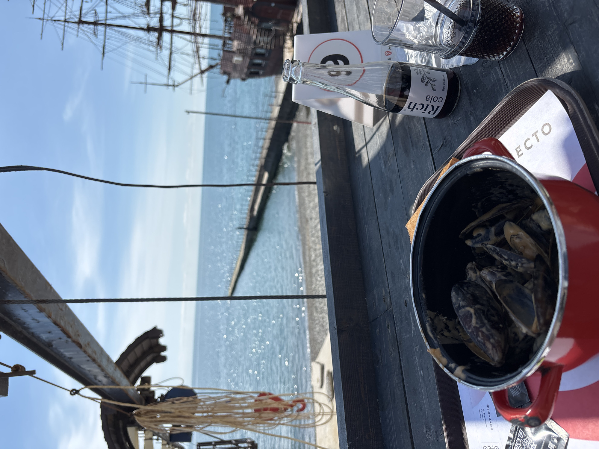 Here is a detailed description of the image:
**Scene:** The image captures a meal setting on a wooden table at an outdoor location. In the foreground is a red pot filled with cooked mussels, placed on a tray with a paper or menu visible underneath. Next to the pot are a glass bottle of "Rich Cola" and two glasses. The background features a view of the sea, a pier, and a distant sailing ship. A partial structure of a boat or platform is visible on the left side of the frame.
**Objects:**
* **Mussels:** Cooked mussels in a red pot.
* **Beverages:** A bottle of "Rich Cola", a glass of dark liquid with a straw, and an empty glass.
* **Table setting:** Wooden table, tray, paper/menu under the pot.
* **Ship:** Distant sailing ship in the background.
**Environment:**
* **Sea:** The scene overlooks a bright blue sea with a visible pier.
* **Location:** The outdoor setting suggests a restaurant or cafe near the coast, possibly in a tourist area.
**Inferences:** The image likely captures a meal being enjoyed at a seaside restaurant or cafe, with a focus on seafood (mussels) and refreshments. The sailing ship adds a scenic element to the overall atmosphere.
**Sochi, Russia Inference:** The beverage "Rich Cola" is a popular drink brand in Russia, suggesting that the location might be in Russia. The coastal setting and visible pier, coupled with the Russian beverage, are consistent with a possible location in Sochi, Russia, a popular resort city on the Black Sea coast.
