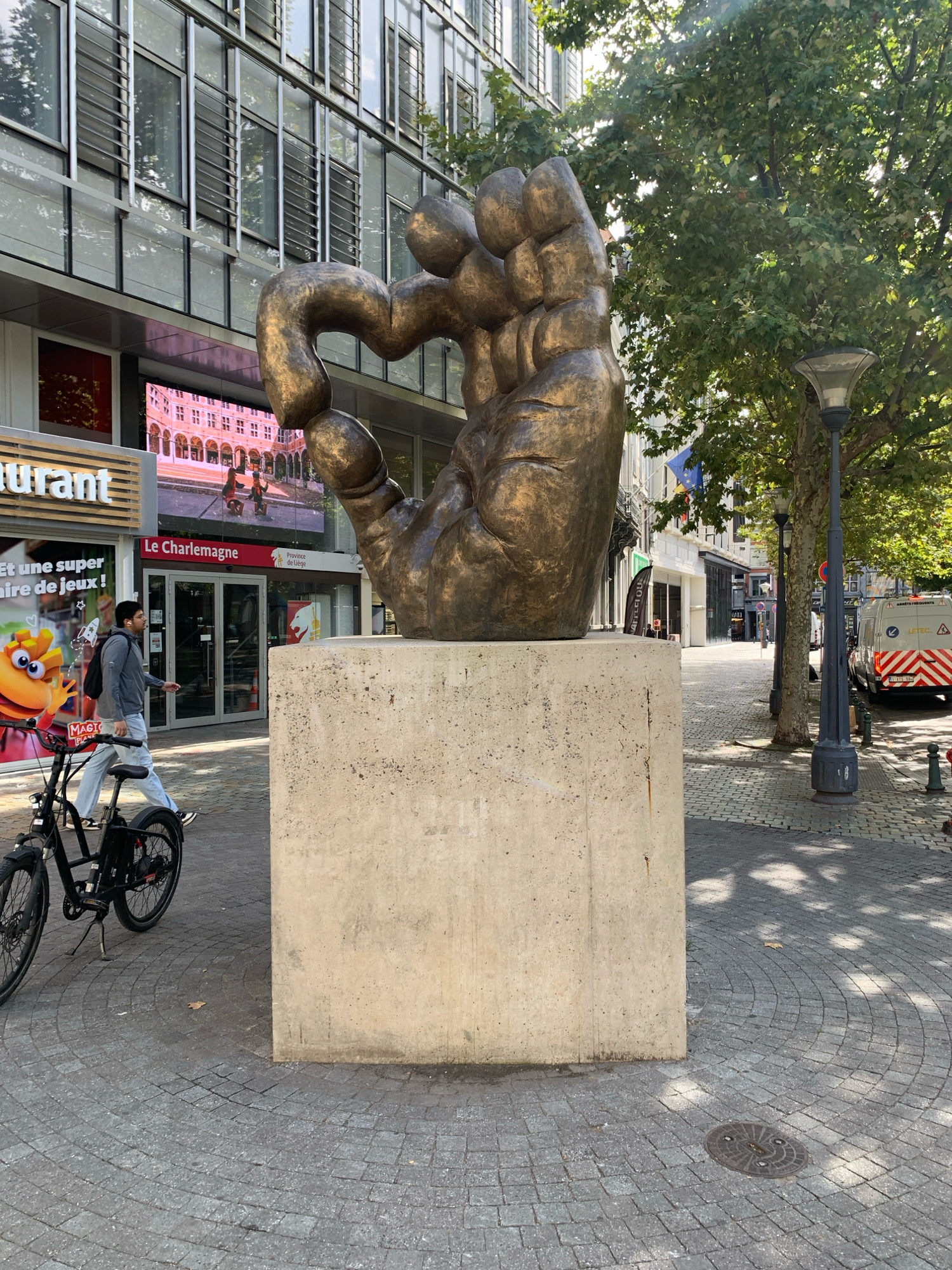 This is a street-level photograph taken in the city of Liège, Belgium. In the foreground, a large, bronze-colored sculpture of a hand is mounted on a concrete pedestal. The sculpture is abstract and appears to be made of interconnected, bulbous forms. To the left of the sculpture, a person is walking their bicycle. The individual is wearing a grey jacket, light-colored pants, and a backpack. The bicycle is black and appears to be an electric bike. The ground is paved with cobblestones arranged in a circular pattern. In the background, a modern building with large windows and reflective surfaces is visible. Below the windows of this building, a digital screen displays an image of a historic building with arches and people dressed in period clothing. A sign above the screen reads "Le Charlemagne" and "Province de Liège". Further down the street, trees line the sidewalk, and a streetlamp is visible. A white service van with orange and white reflective stripes is parked on the right side of the street. The sky is blue and sunny, with dappled sunlight filtering through the trees.