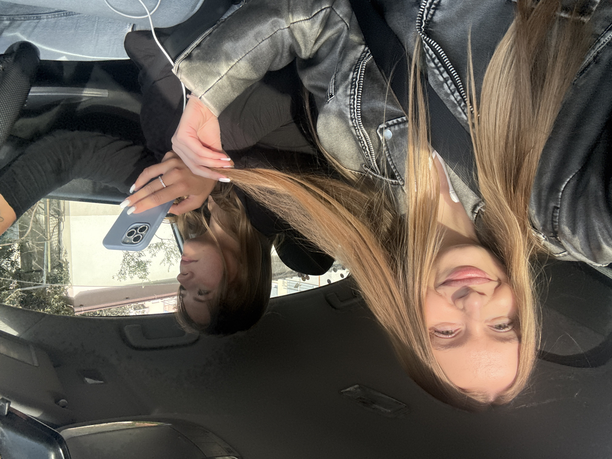 The image is a selfie taken from inside a car, likely by the passenger. The subject is a young woman with long, blonde hair and fair skin, wearing a grey leather jacket. She is holding a smartphone in her right hand, which is visible in the reflection on the car's ceiling. She is wearing a ring on her left hand. The car's interior is visible, including the dashboard and ceiling. Outside the car, the reflection shows a building with white walls and a pink roof, suggesting a residential or commercial building in an urban area. The reflection also shows trees. Considering the description of the city of Budva, Montenegro, known for its architecture and coastal views, the location could plausibly be within the city limits, but without further visual context (such as street signs or distinct landmarks) pinpointing the exact location is impossible.