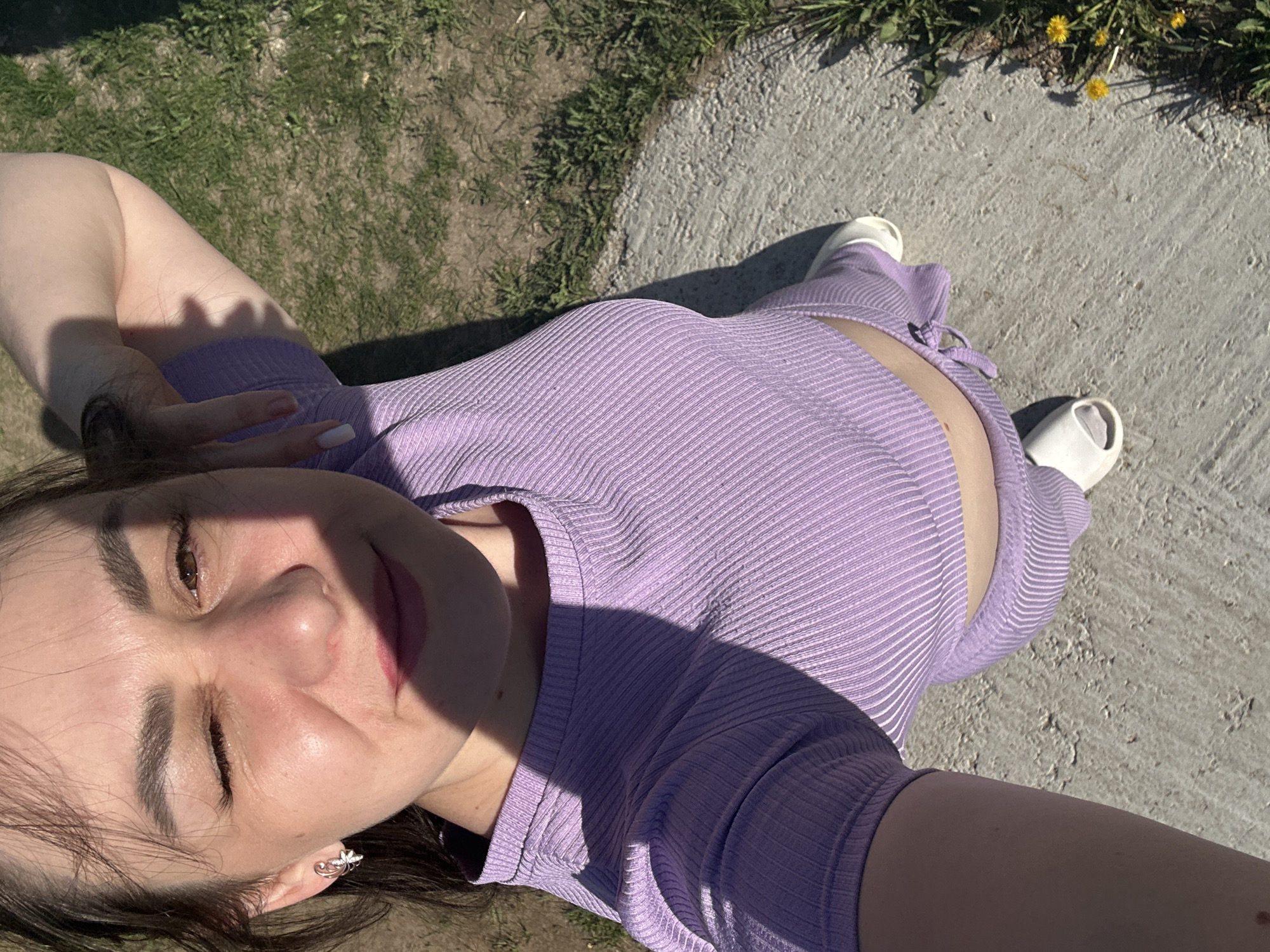 A woman is lying on a concrete surface next to a patch of grass and dandelions. She is wearing a light purple ribbed top and pants and white slip-on shoes. She appears to be taking a selfie in bright sunlight, with part of her face in shadow. It is not possible to confirm the exact location within Chaikovsky, Russia from the image alone.