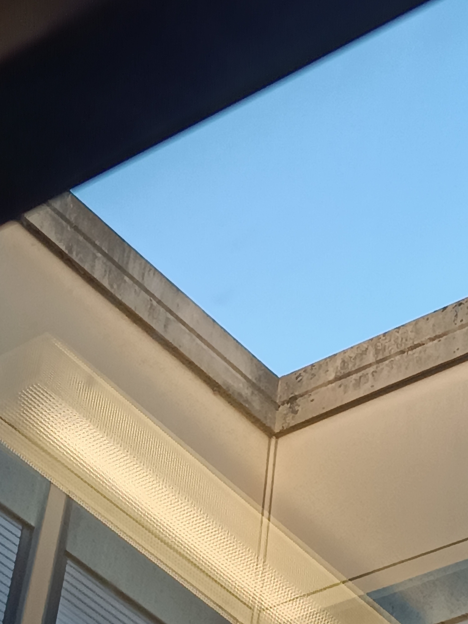 The image shows a portion of a skylight with a clear blue sky visible above. The concrete framing of the skylight is weathered and shows signs of age. Below the skylight, a textured glass panel with a ribbed pattern is visible, suggesting it is part of a window or a diffuser. The surrounding interior architecture appears to be a light-colored, smooth surface. The perspective suggests the viewer is looking upwards from inside a building. There are no people or specific activities depicted. The scene is indoors looking out through a skylight. The location in Casalmaggiore, Italy, is not discernible from the visual information in the image.