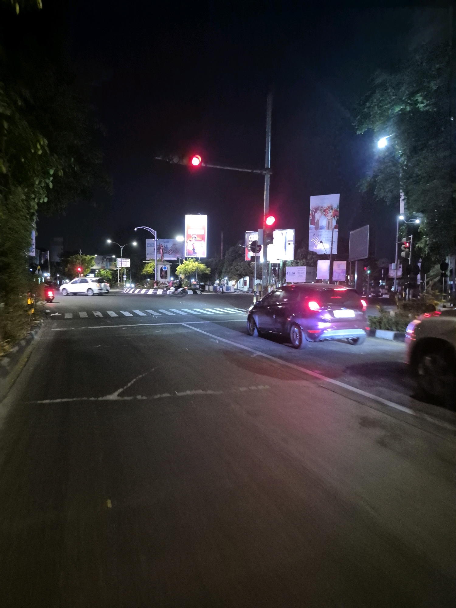 The image depicts a street scene at night in Surakarta, Indonesia. The view is from a low angle, looking down a road that appears to be an intersection with a roundabout. Traffic lights are visible and illuminated with red signals, indicating that vehicles should stop. A purple hatchback car is in the foreground, its brake lights reflecting on the wet asphalt. To the left, a white SUV is stopped at the intersection, also under a red light. A red scooter is visible to the far left, stopped near a tree-lined sidewalk. Several large illuminated billboards are visible in the background, displaying various advertisements. The sky is dark, and the overall lighting suggests it is nighttime. Trees and streetlights are also present, contributing to the urban nocturnal ambiance.