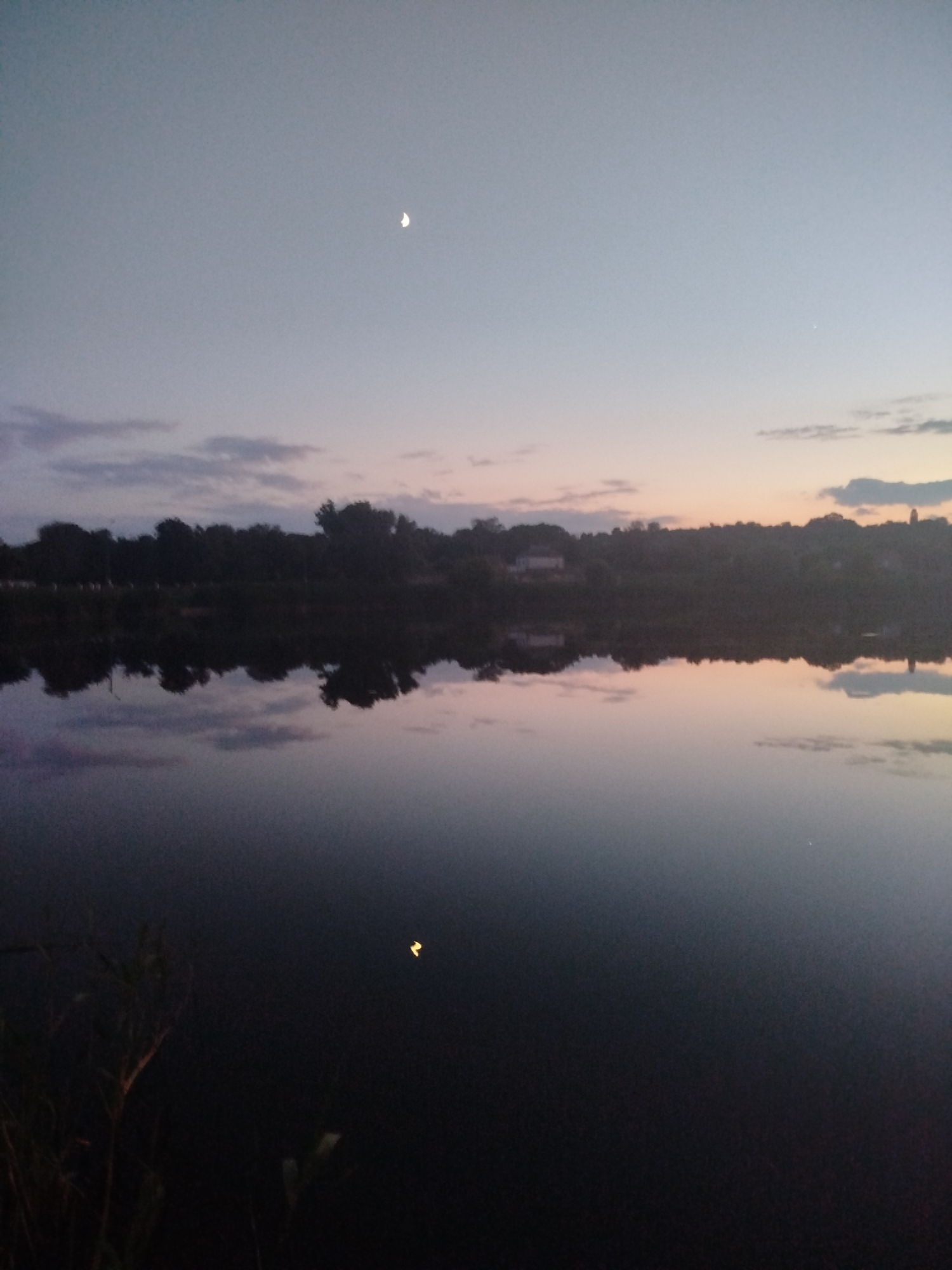This image depicts a body of water at dusk, likely a lake or river, reflecting the twilight sky and a silhouetted treeline. In the upper portion of the sky, a crescent moon is visible. The water's surface is calm, creating a mirror-like reflection of the sky and the trees along the shore. The overall atmosphere is serene and peaceful. This scene could be located in Ustyvytsia, Ukraine, given the context provided.