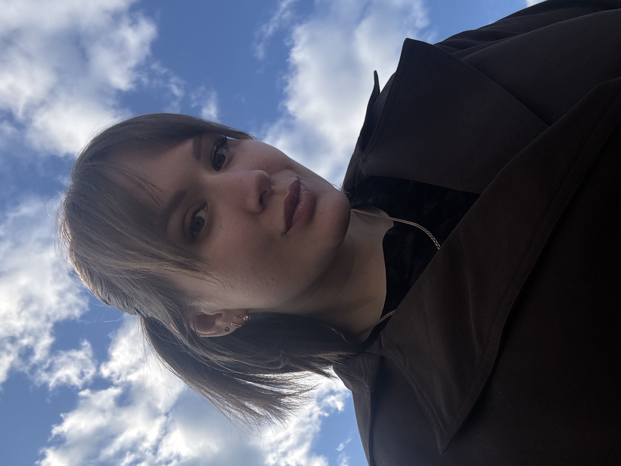 A selfie-style, low-angle shot shows a person with light skin and brown hair posing against a backdrop of a blue sky with scattered white clouds. The person has a side-swept fringe and their hair is partially tied back. They are wearing a dark brown jacket and a black top underneath, with a thin gold chain visible around their neck. The photograph's location is not definitively identifiable as Chaikovsky, Russia from the visual information alone.
