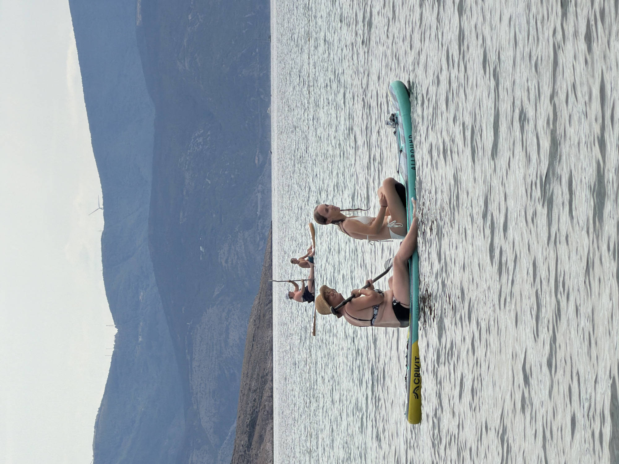 Two women are paddleboarding on the water. The woman in the foreground on the left is wearing a straw hat and a black and white patterned bikini. She is sitting on a yellow and teal paddleboard with the brand "CRIVIT" visible. The woman in the foreground on the right is wearing a white bikini. She is sitting on a teal paddleboard with "ALLROUND" visible. In the background, two other individuals are paddleboarding further away on their boards. The scene is set in a body of water with a hilly, mountainous coastline in the distance, characteristic of Općina Baška, Croatia. The sky is overcast.