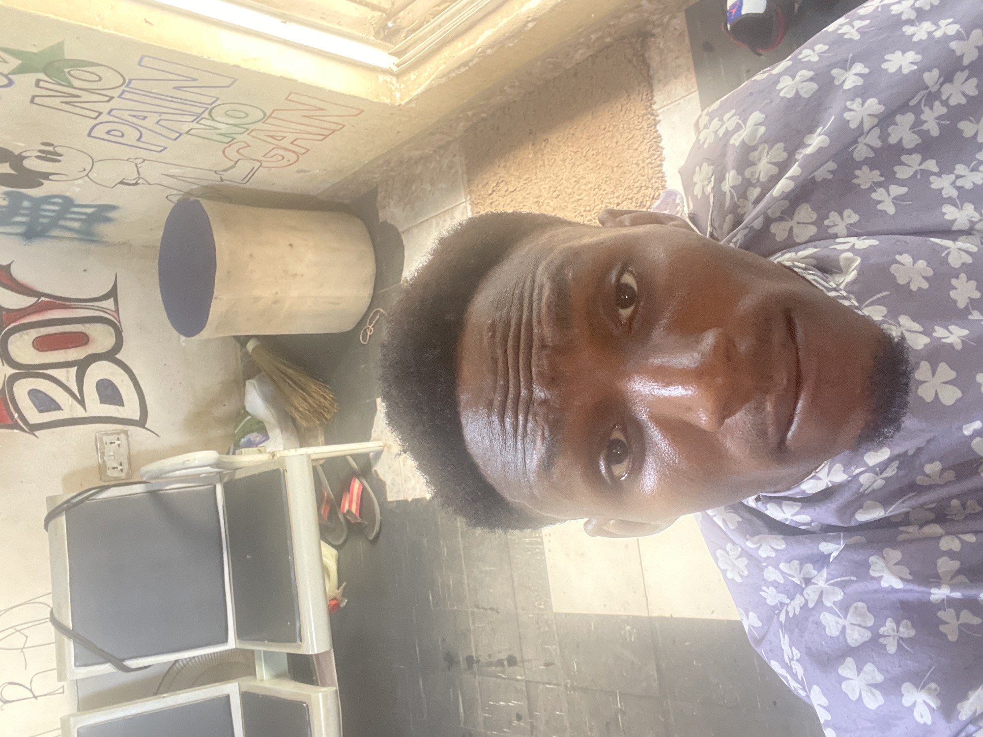 A man is taking a selfie in a barber shop in Akowonjo, Nigeria. He is wearing a light purple shirt with white clover patterns and a darker purple cape around his neck. The background shows two dark-colored barber chairs, a cylindrical stool, a broom, and wall art that includes the words "BOY" and "NO PAIN NO GAIN", along with a cartoon character that resembles Mickey Mouse. There are also some flip-flops visible on the floor beneath the chairs. The lighting suggests the image was taken during the day, with natural light coming from a window on the right side of the frame.