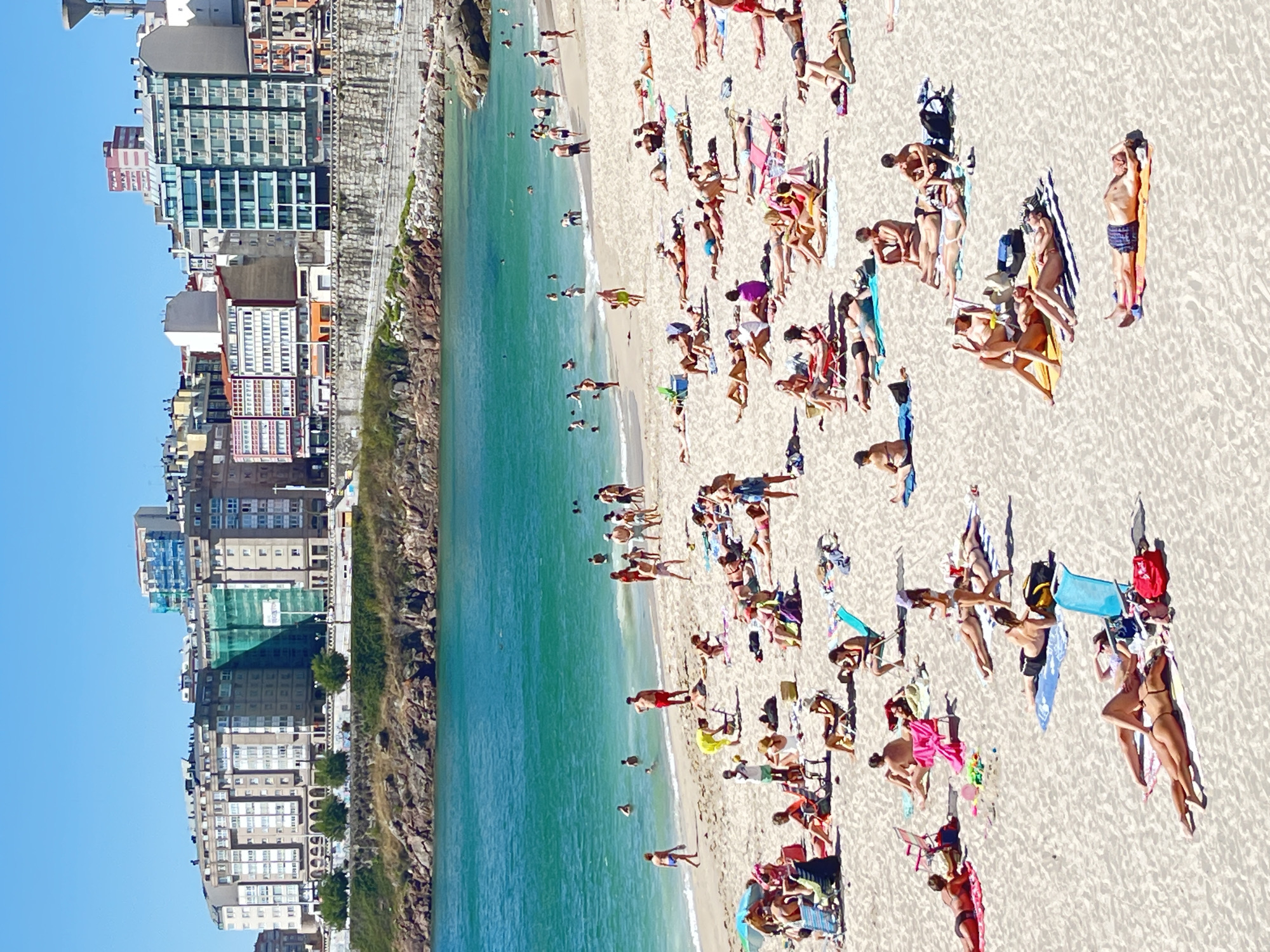 This is an aerial, vertical, panoramic shot of a crowded beach in A Coruña, Spain, on a sunny day. The beach is populated with many people sunbathing, swimming, and relaxing. They are spread across the sand, some lying on towels, others on beach chairs or directly on the sand. Several are in the ocean, enjoying the turquoise water. The setting features a backdrop of tall, modern buildings and a rocky coastline on the left side of the image, with the clear blue sky above. The sun casts distinct shadows, indicating it is likely midday. The overall atmosphere is one of leisure and enjoyment. No visible text is present in the image.