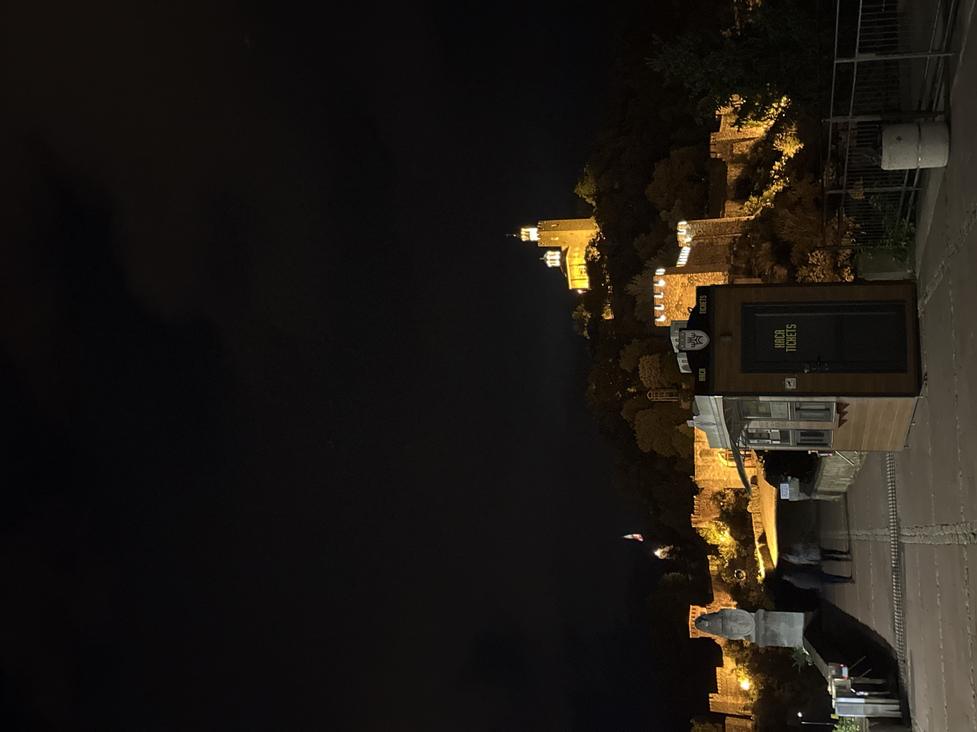 Captured at night, the image showcases the illuminated medieval fortress of Tsarevets in Veliko Tarnovo, Bulgaria, seen from across a dark void, likely the Yantra River valley. The fortress walls are outlined in light, contrasting against the darkness. On the right side, there's a paved walkway with a ticket booth labeled "Tsarevets Tickets." A couple of figures, potentially visitors, are walking on the path near the booth. Overhead structures and railings are visible along the pathway. The overall scene exhibits the historical architecture lit up in the darkness of the Bulgarian night.